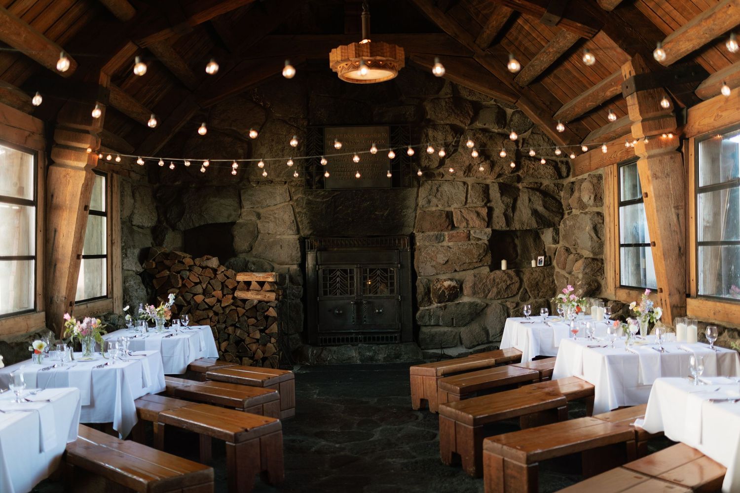Rustic stone wedding reception hall with string lights, wood benches, and floral centerpieces.