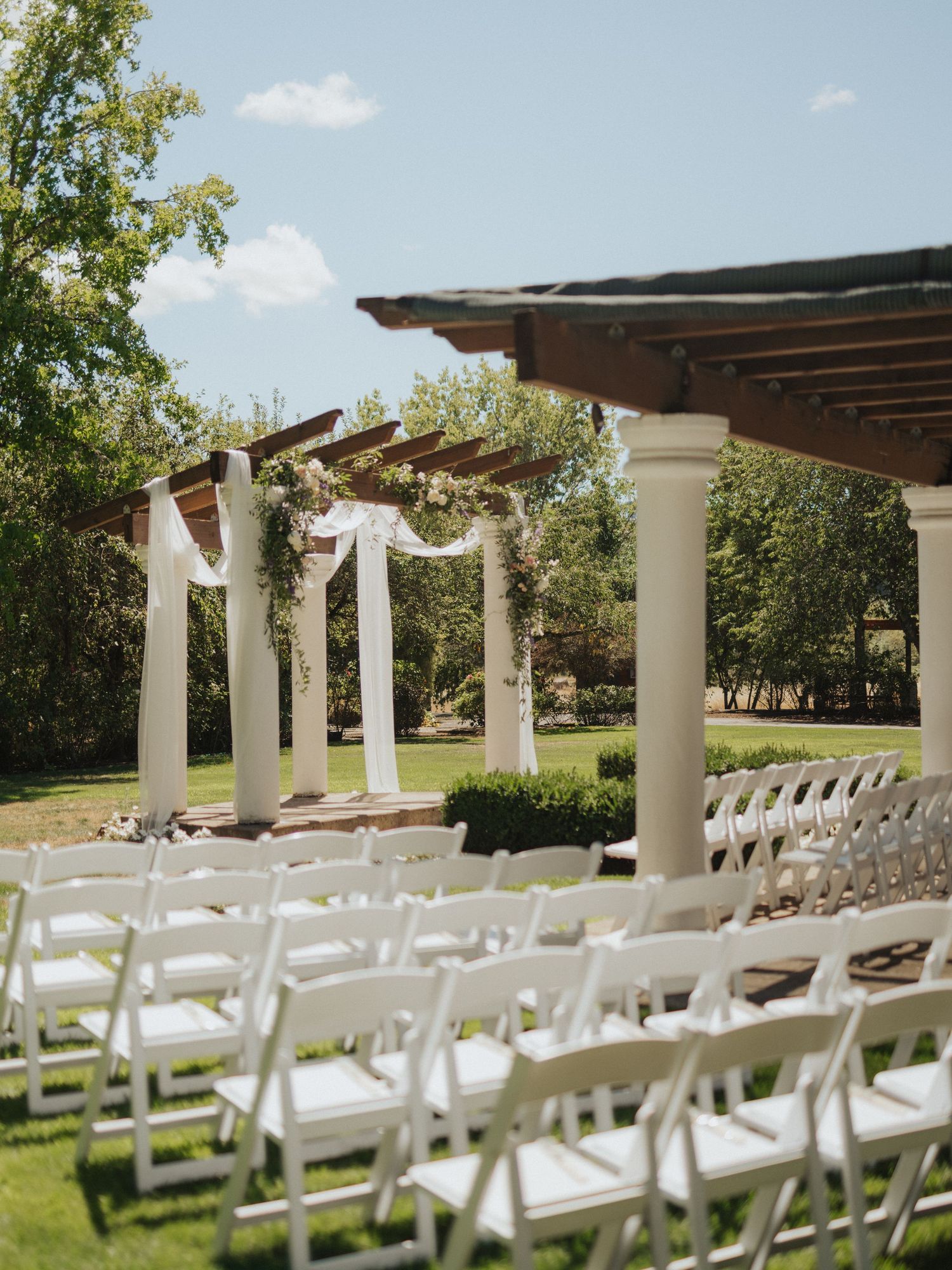 Outdoor wedding ceremony setup with white folding chairs and floral-draped pergola on a sunny summer day.
