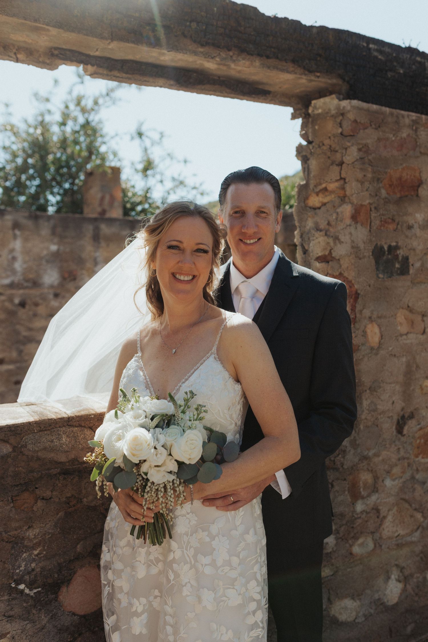 Smiling bride and groom pose with white bouquet against rustic stone wall in golden sunlight.