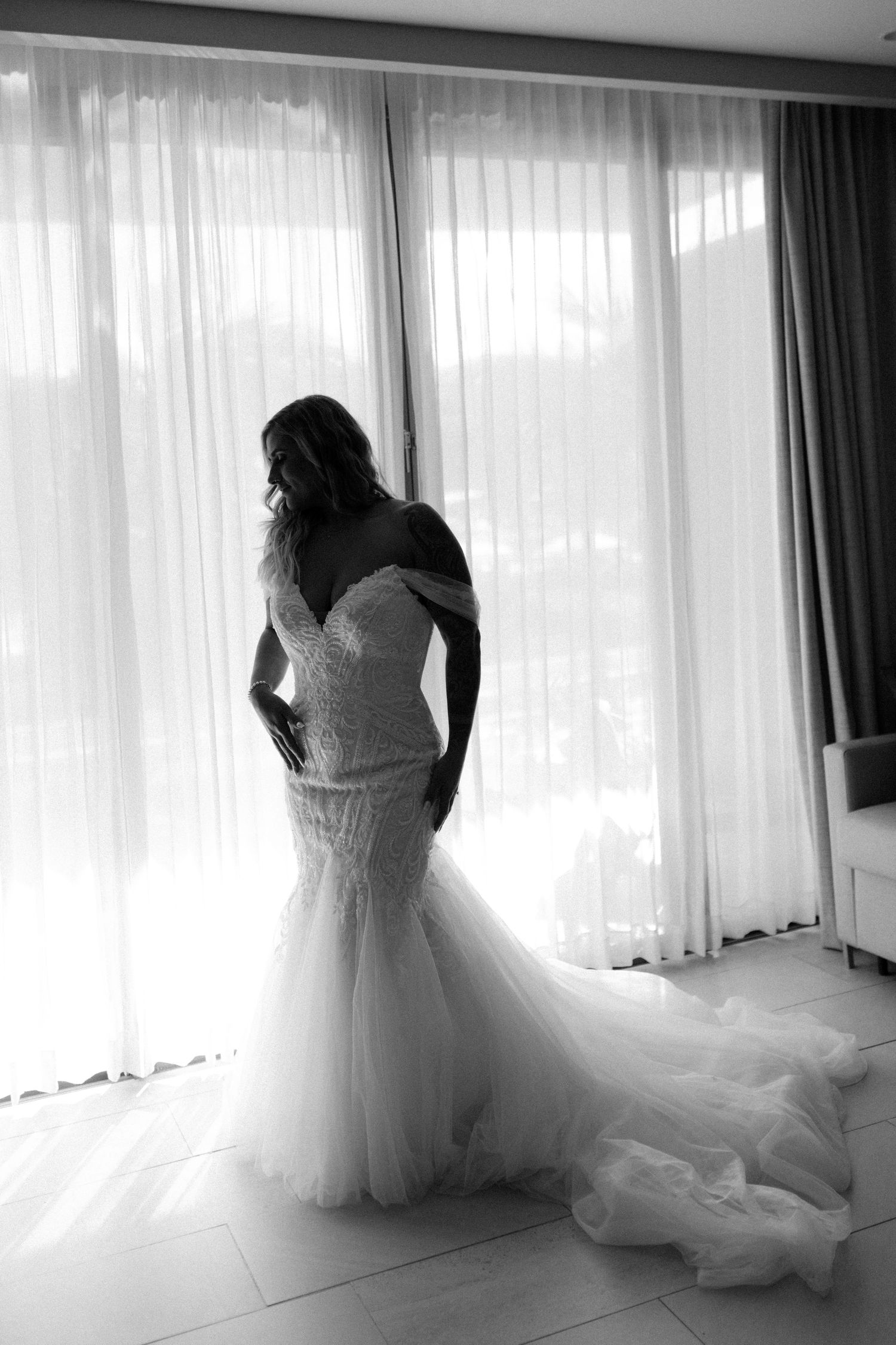 Elegant bride in mermaid wedding gown with long train silhouetted against sheer curtained window.