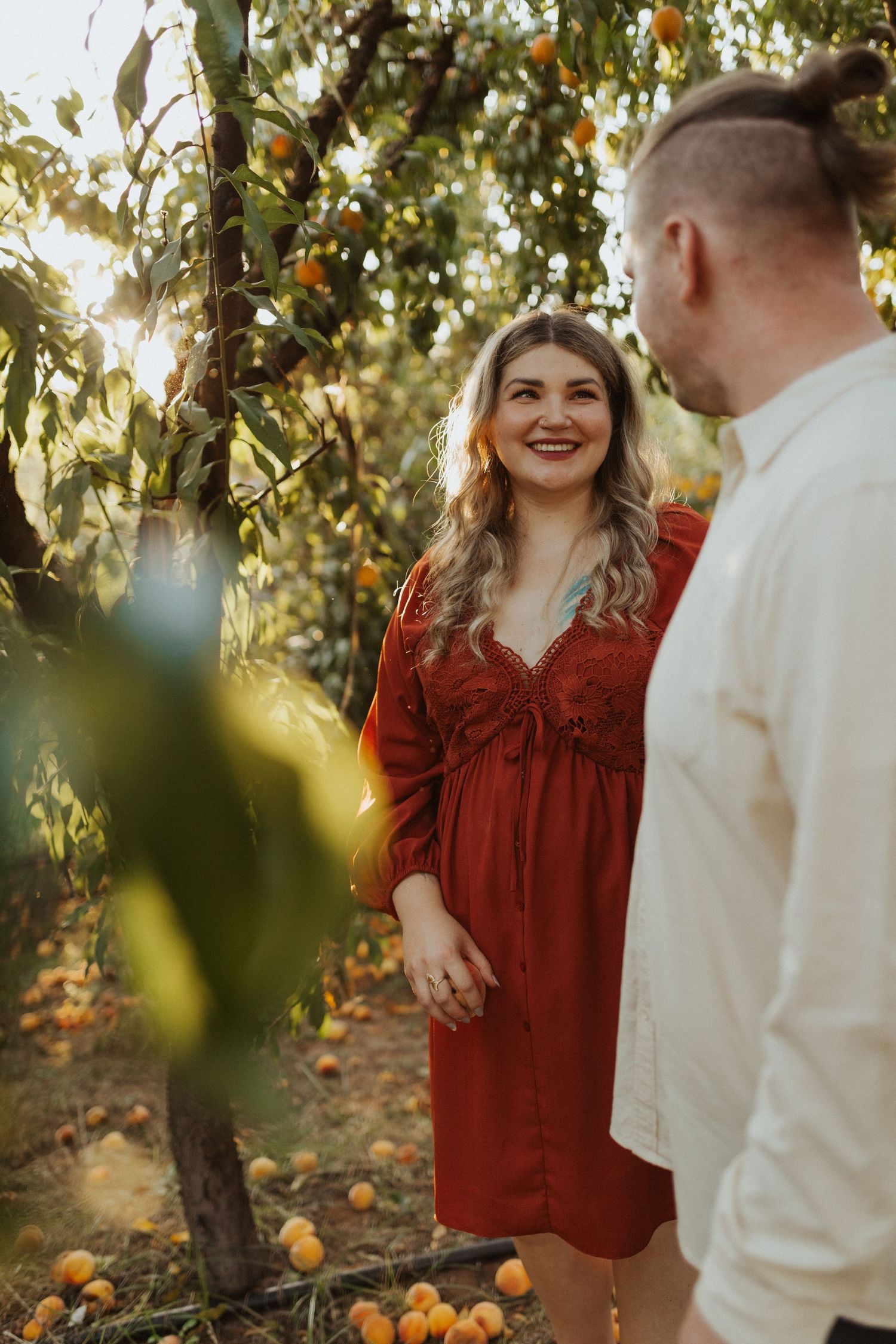 Smiling pregnant woman in rust red dress holds mans hand in sunny orchard with ripe fruit trees.