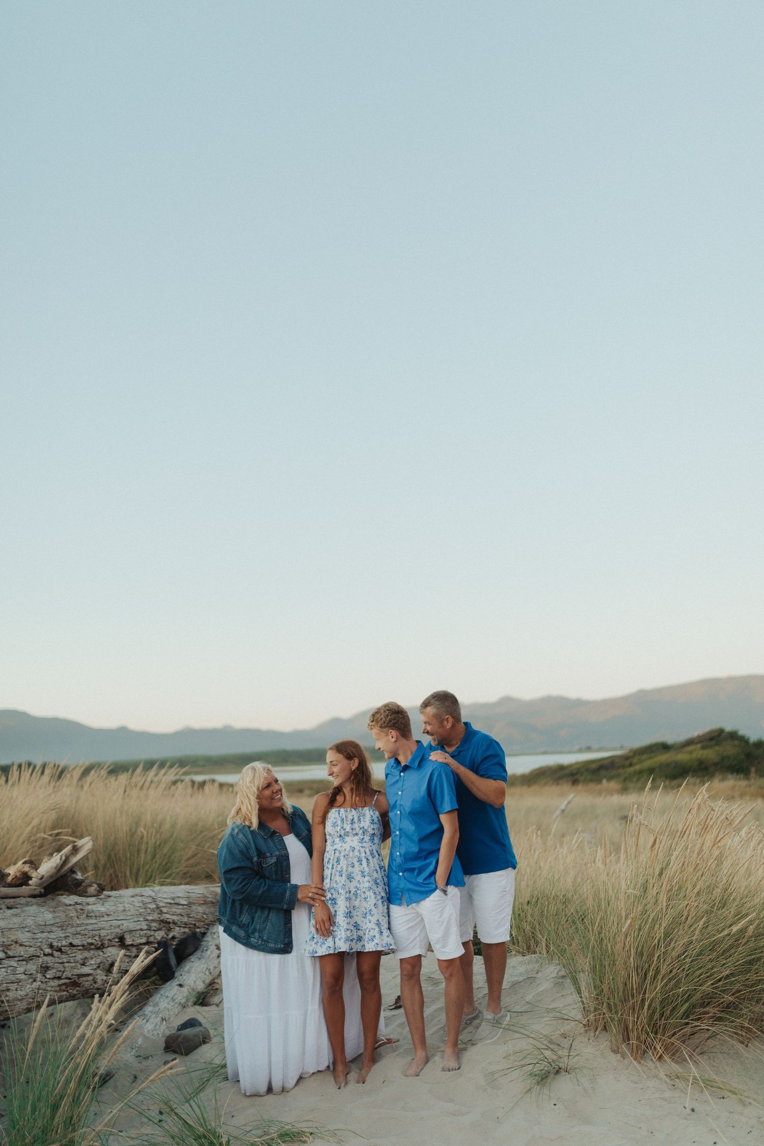 Family of four sharing a candid moment on sandy beach dunes surrounded by tall coastal grass.
