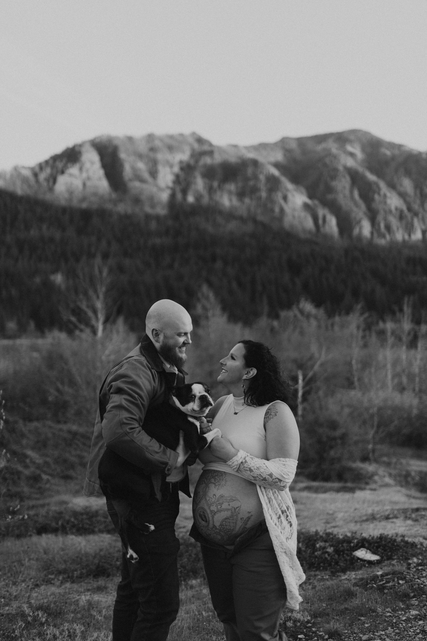 Black and white maternity photo of couple holding their dog while gazing at each other with mountains behind.
