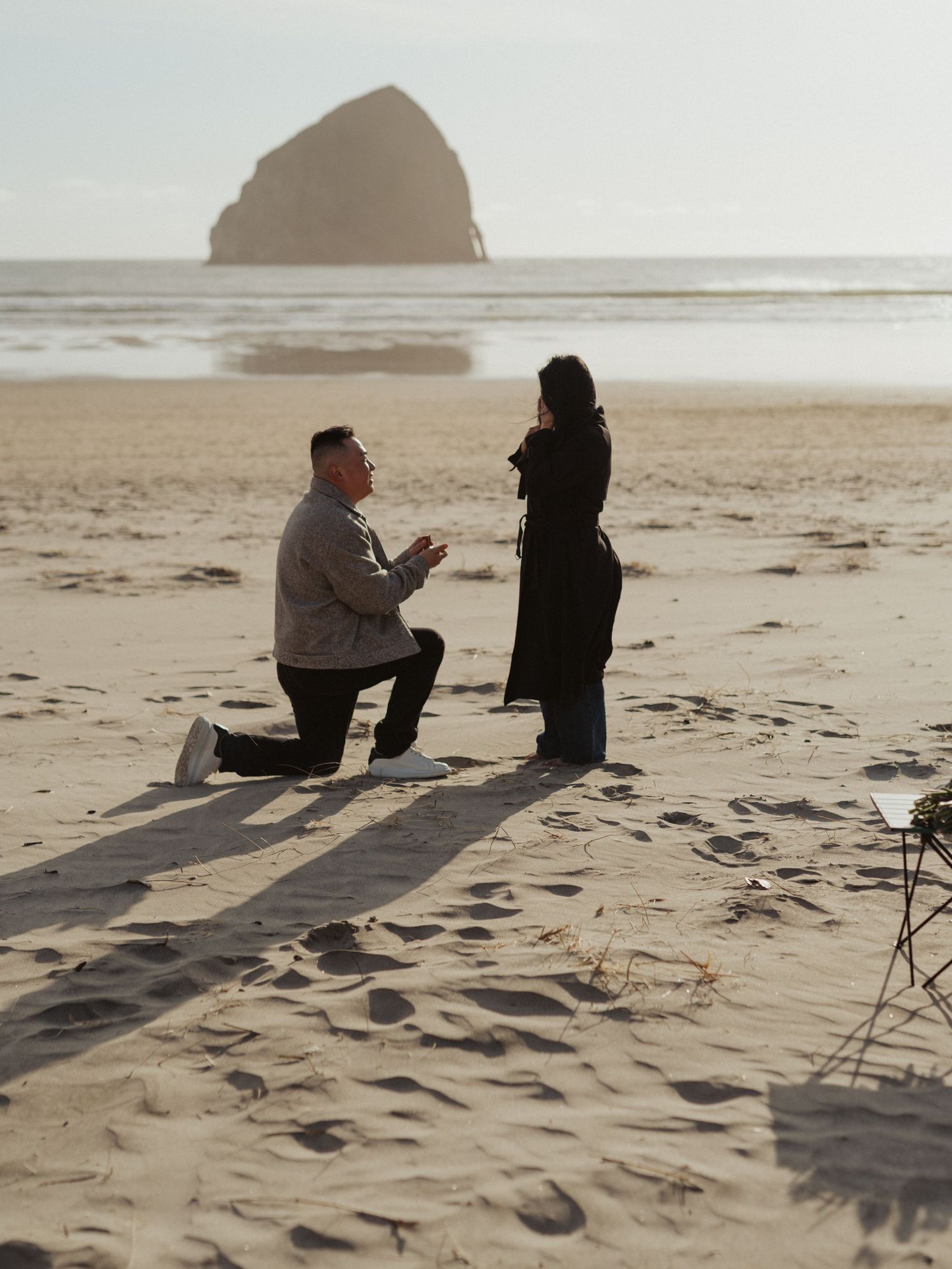 Man proposing on one knee to woman on sandy beach with iconic Haystack Rock in background.