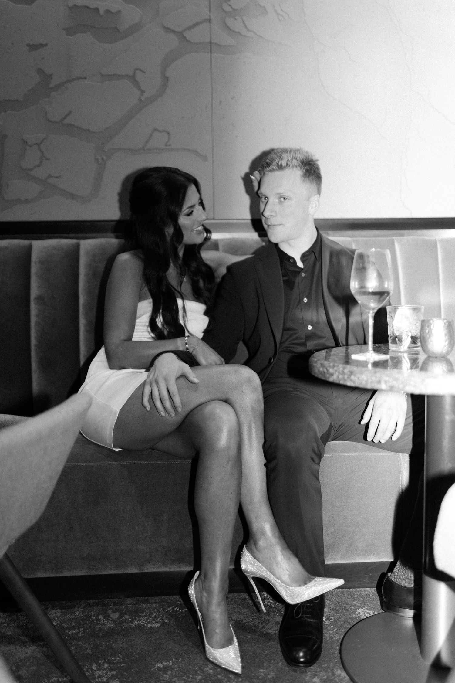 Black and white photo of stylish couple seated at restaurant table with wine, gazing at each other intimately.