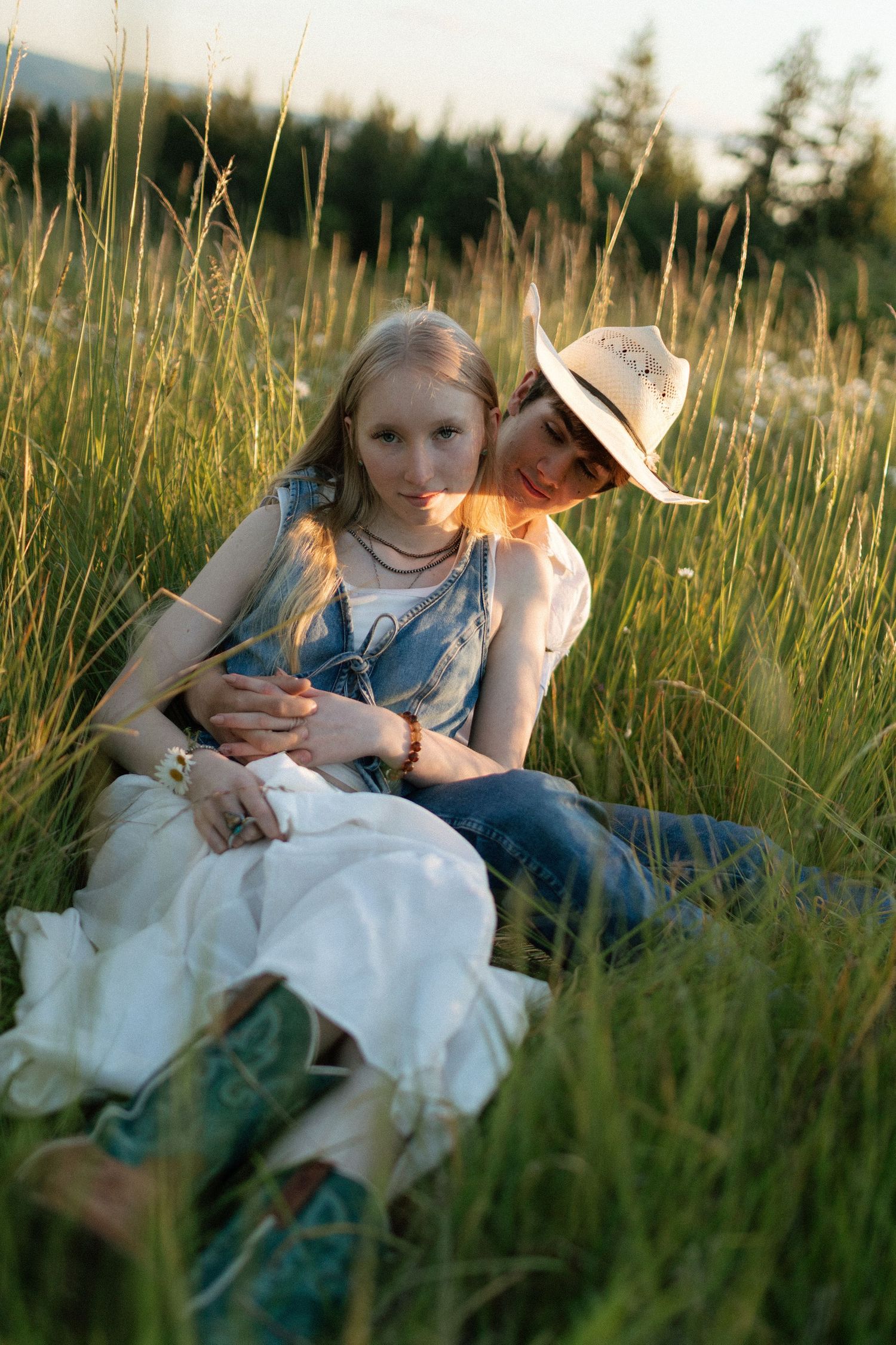 Couple sitting together in tall grass at golden hour, woman in white dress leaning against man in cowboy hat.