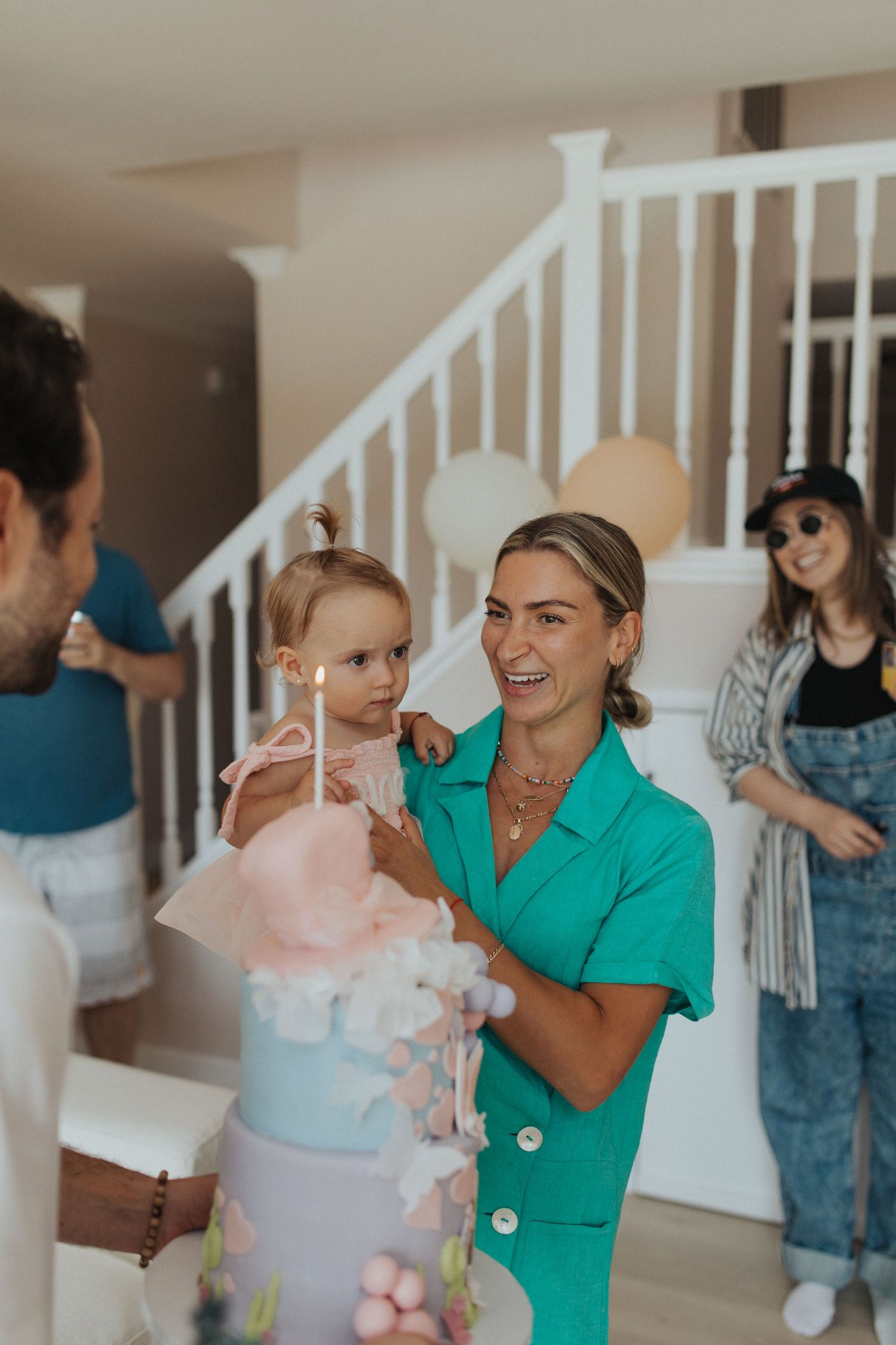 Smiling mother in green dress holds toddler near first birthday cake with lit candle indoors.