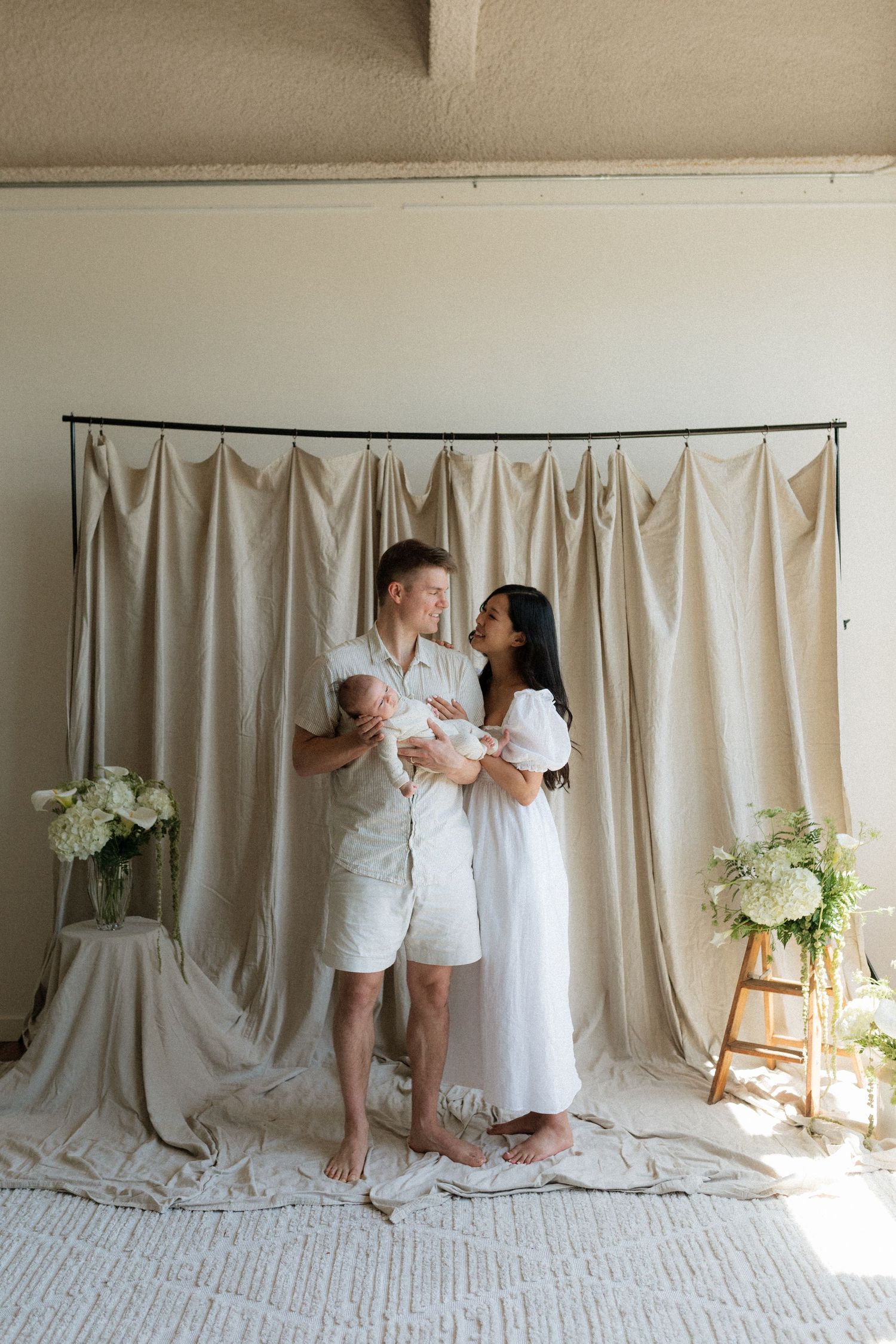 A couple stands barefoot holding their newborn baby in front of a neutral linen backdrop.