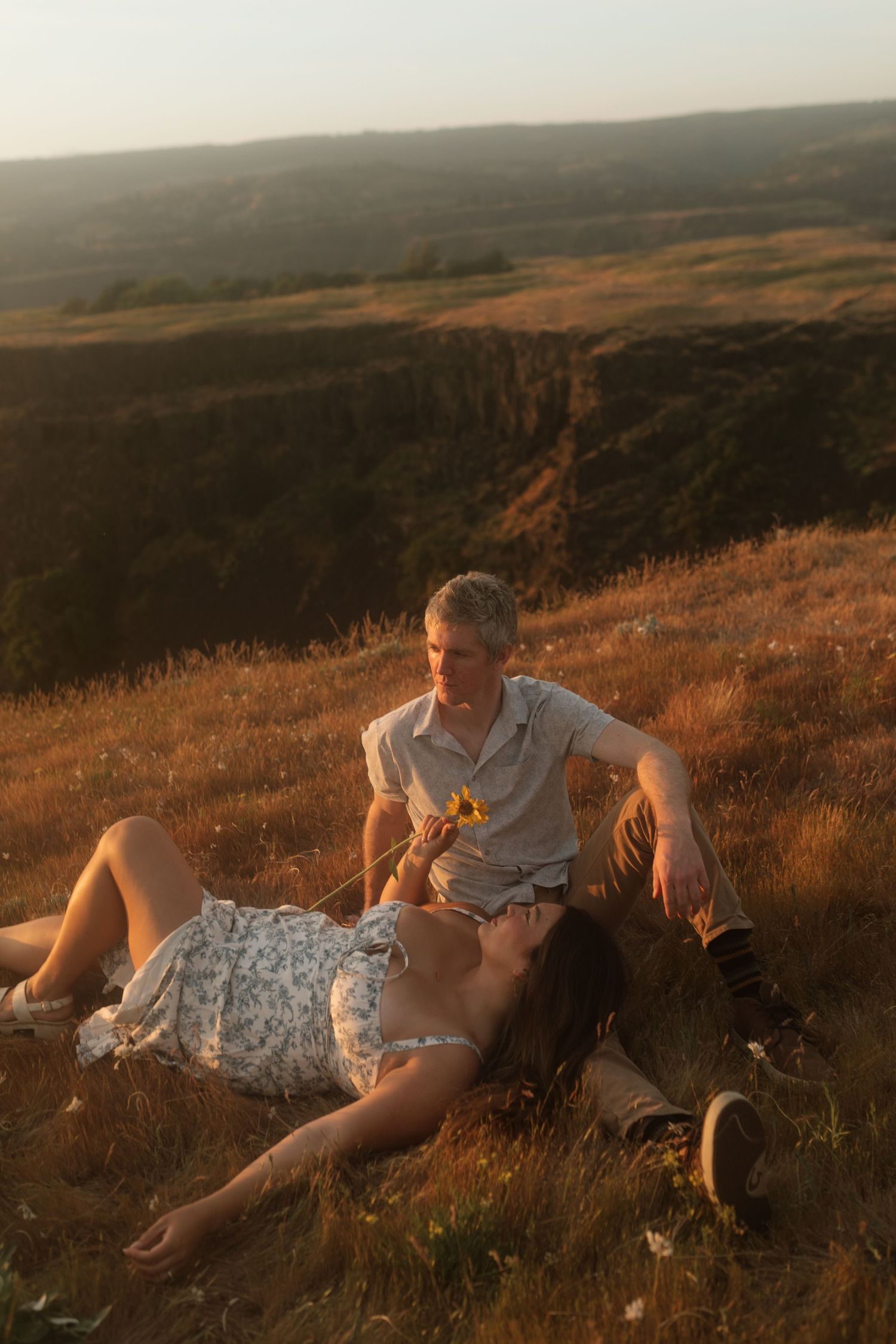 Couple relaxing on golden hillside at sunset, woman lying in grass while man sits beside her holding a wildflower.