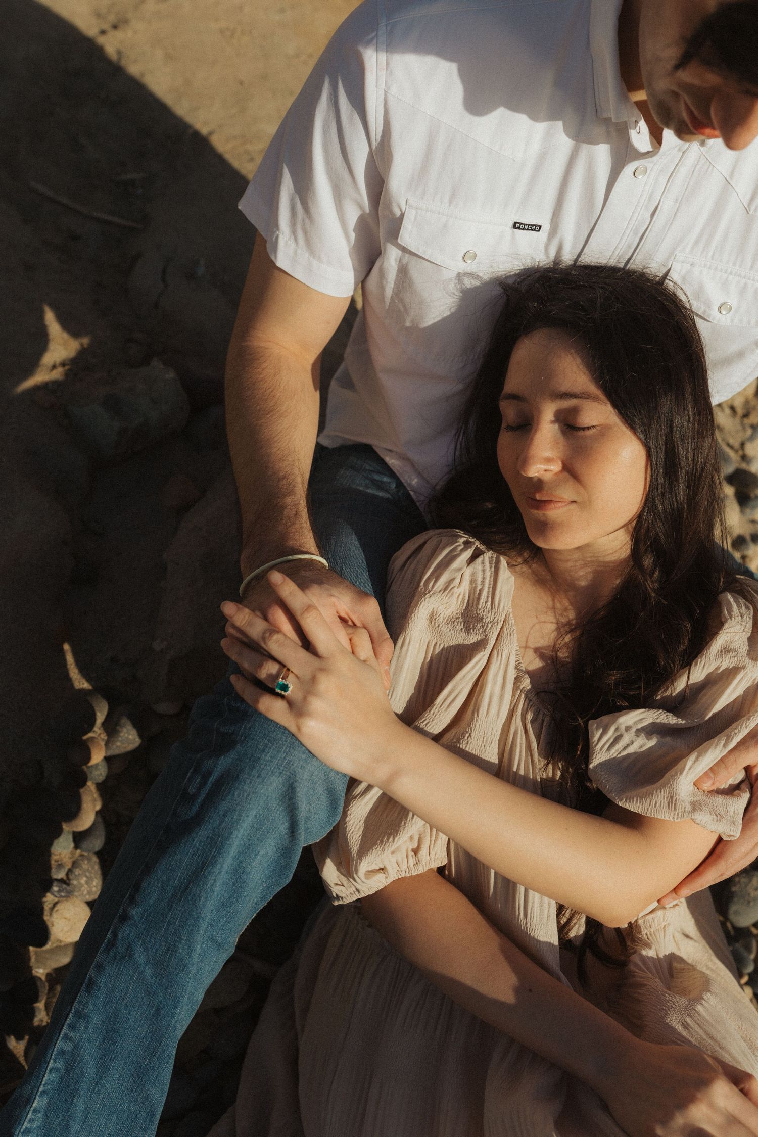 Couple holding hands on rocky beach, woman in beige dress leaning against man in white shirt and jeans.