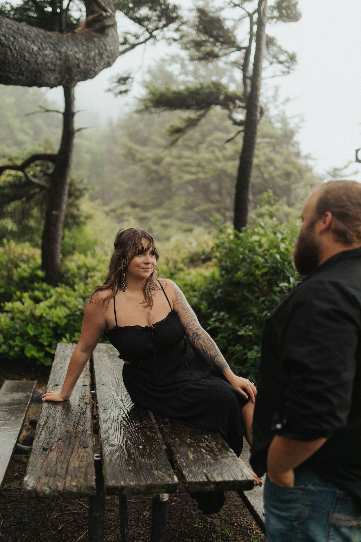 Woman in black dress with tattoos sits on wet picnic table in misty forest during couples photo session.