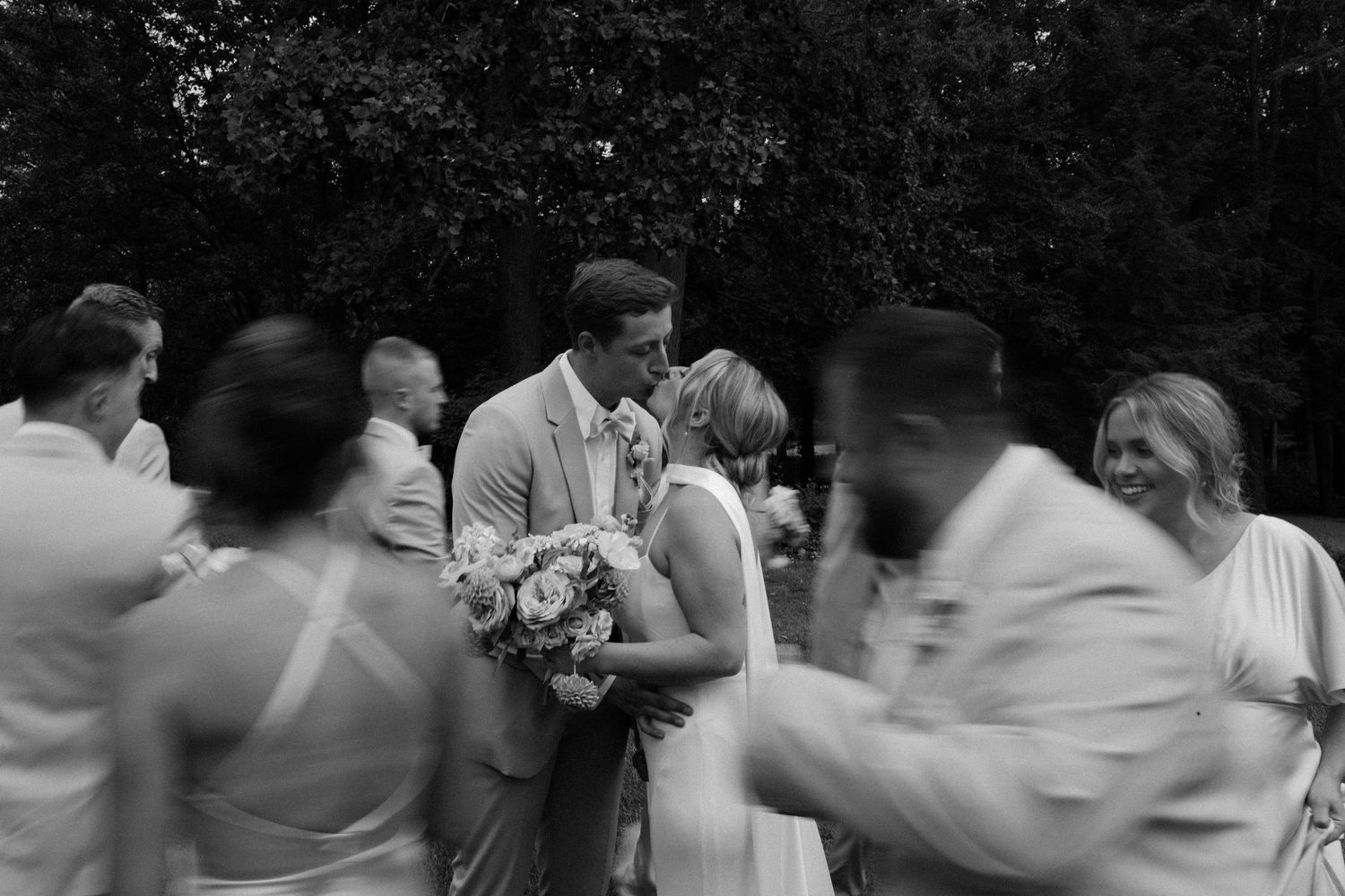 Black and white photo of newlyweds sharing first kiss surrounded by celebrating wedding guests.