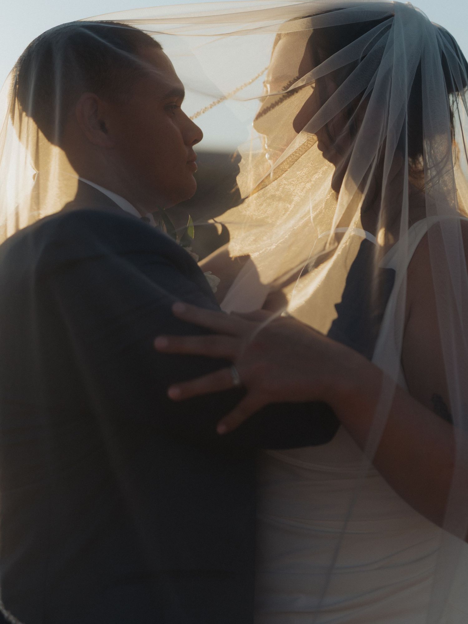 Romantic couple embraces beneath a flowing bridal veil with warm golden backlight creating a dreamy effect.