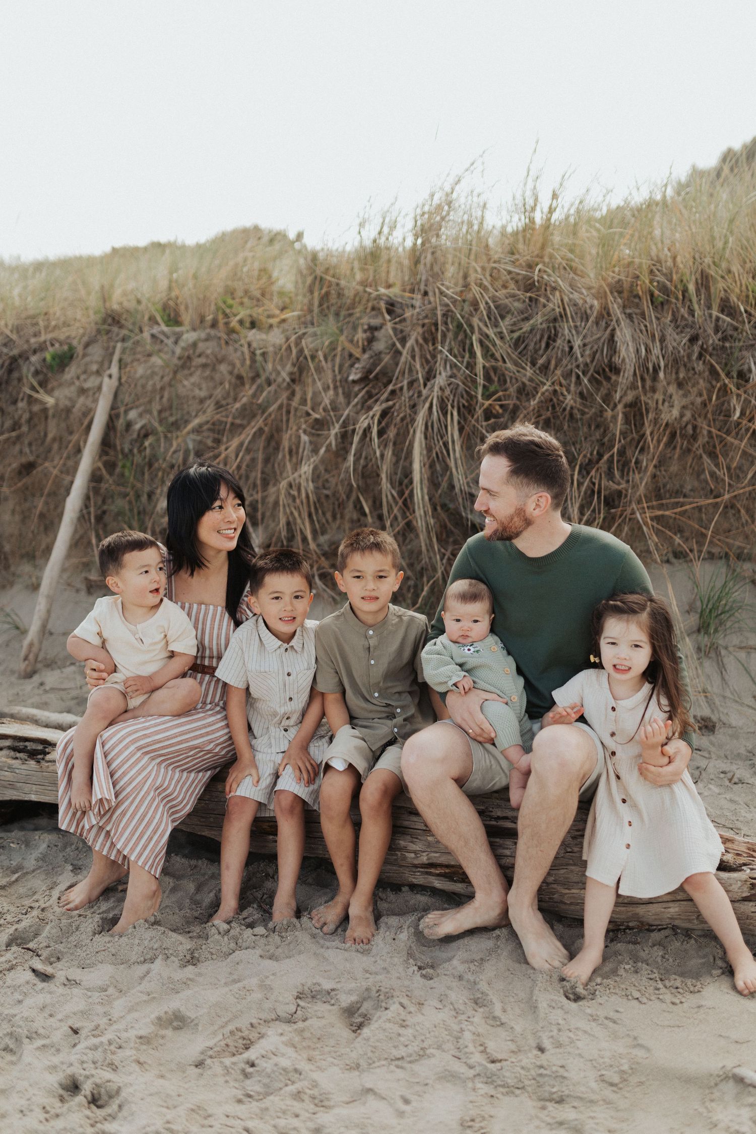Family of seven with five young children sitting together on driftwood at a sandy beach.