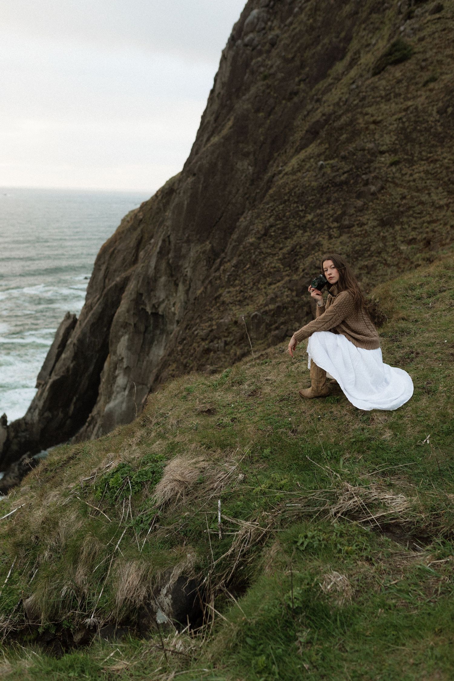 A woman in a brown sweater and white skirt crouches on a grassy coastal cliff holding a film camera.