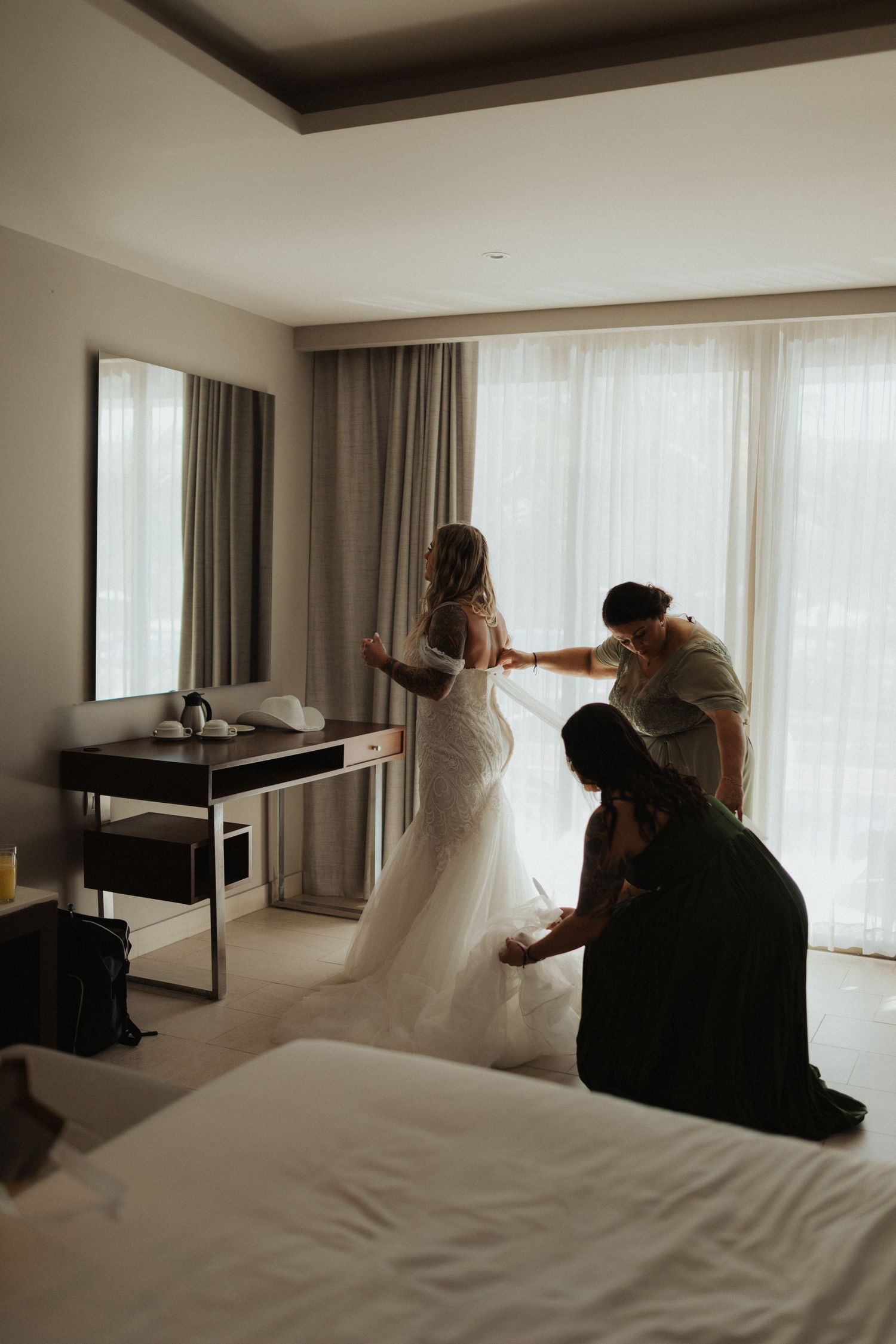 Bride in fitted lace wedding dress being helped by two bridesmaids in a bright hotel room before the ceremony.
