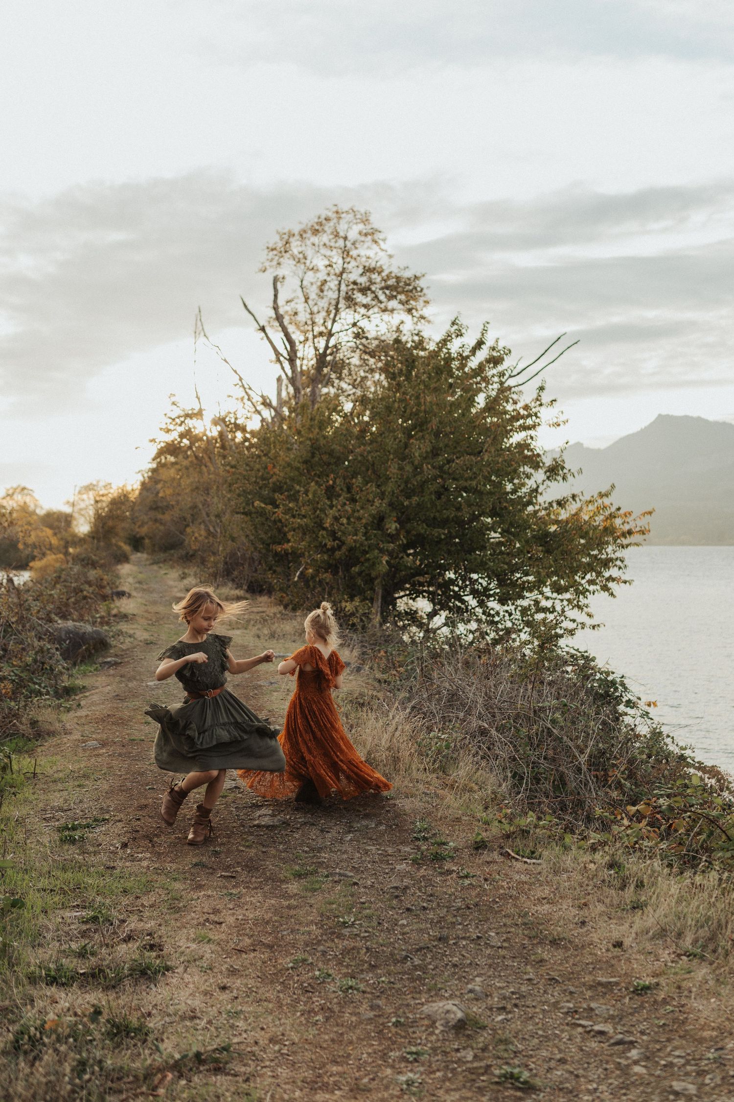 Two young girls in colorful dresses dancing on scenic lakeside path at golden hour sunset.
