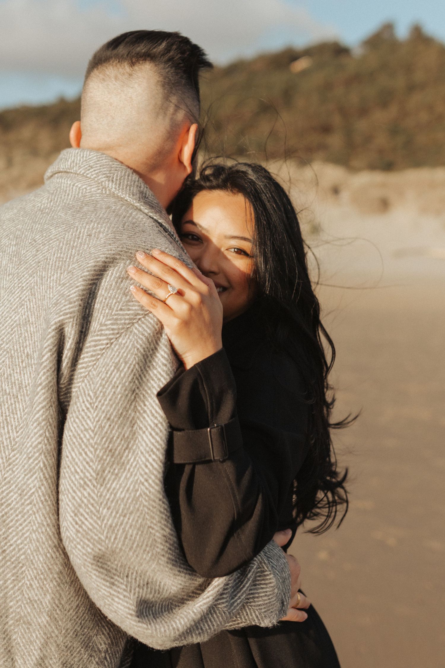 Woman with long dark hair smiles while embracing man on sunny beach, showing off engagement ring.