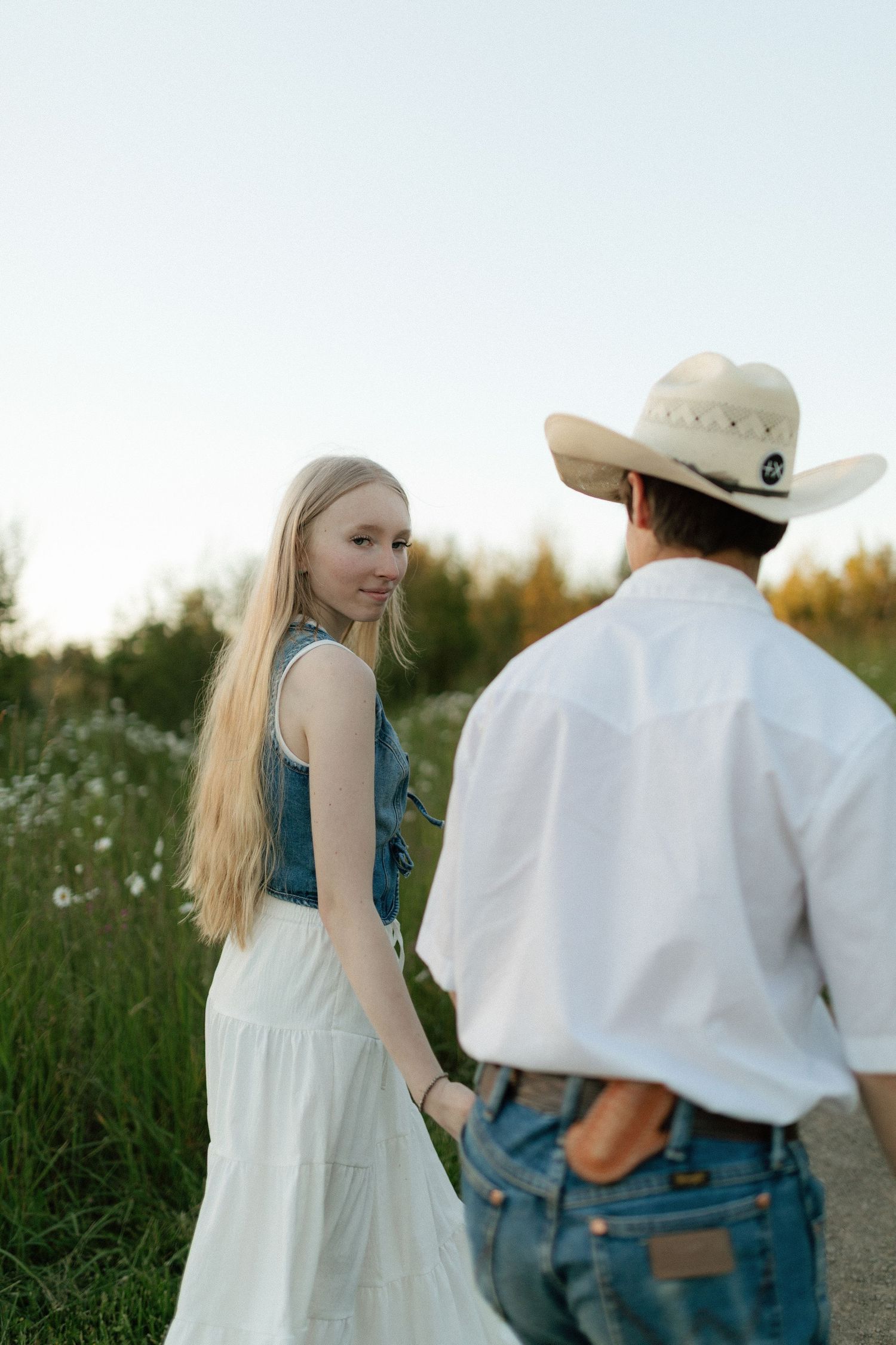 Young woman in white dress glancing back while holding hands with man in cowboy hat on rural path.