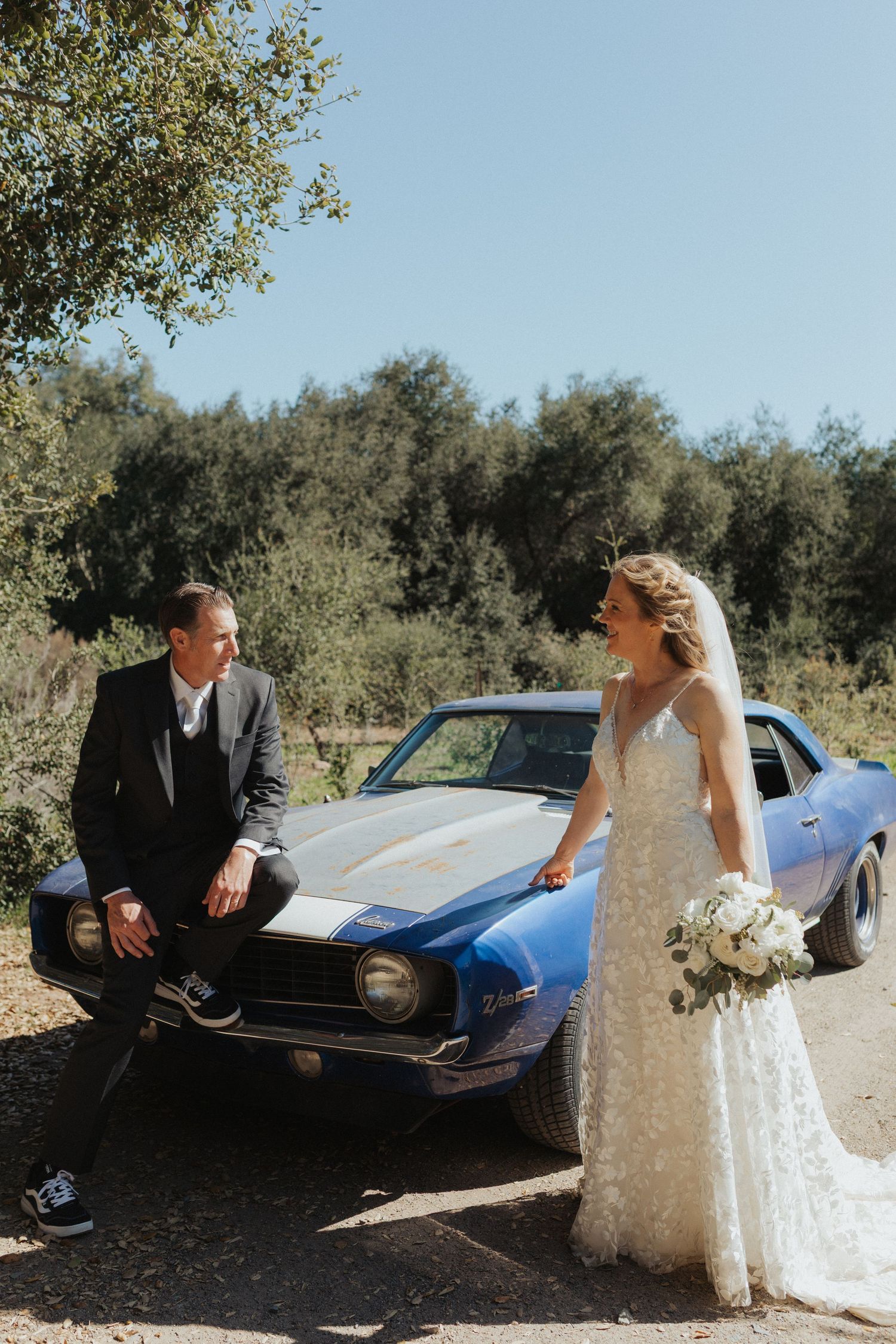 Bride and groom laugh together leaning against a vintage blue muscle car outdoors.