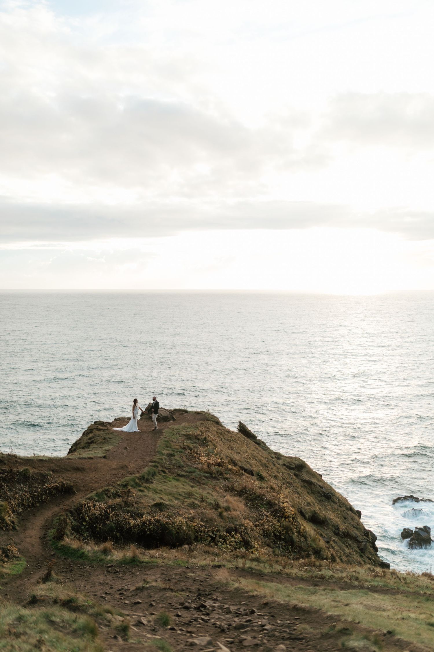 Bride and groom walking along a narrow coastal headland overlooking the ocean.