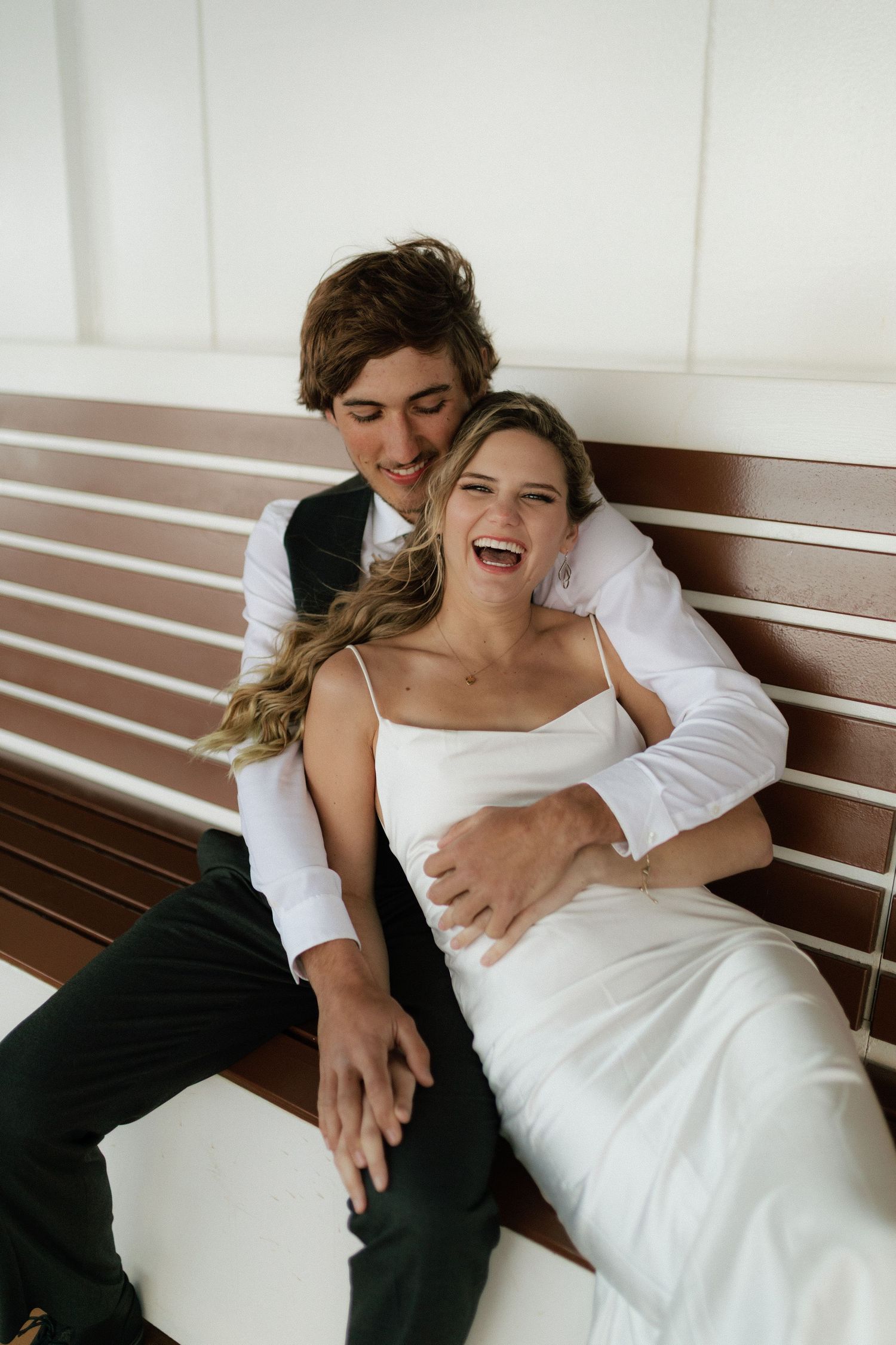 Laughing bride in white dress sits with groom on a wooden bench aboard a cruise ship.