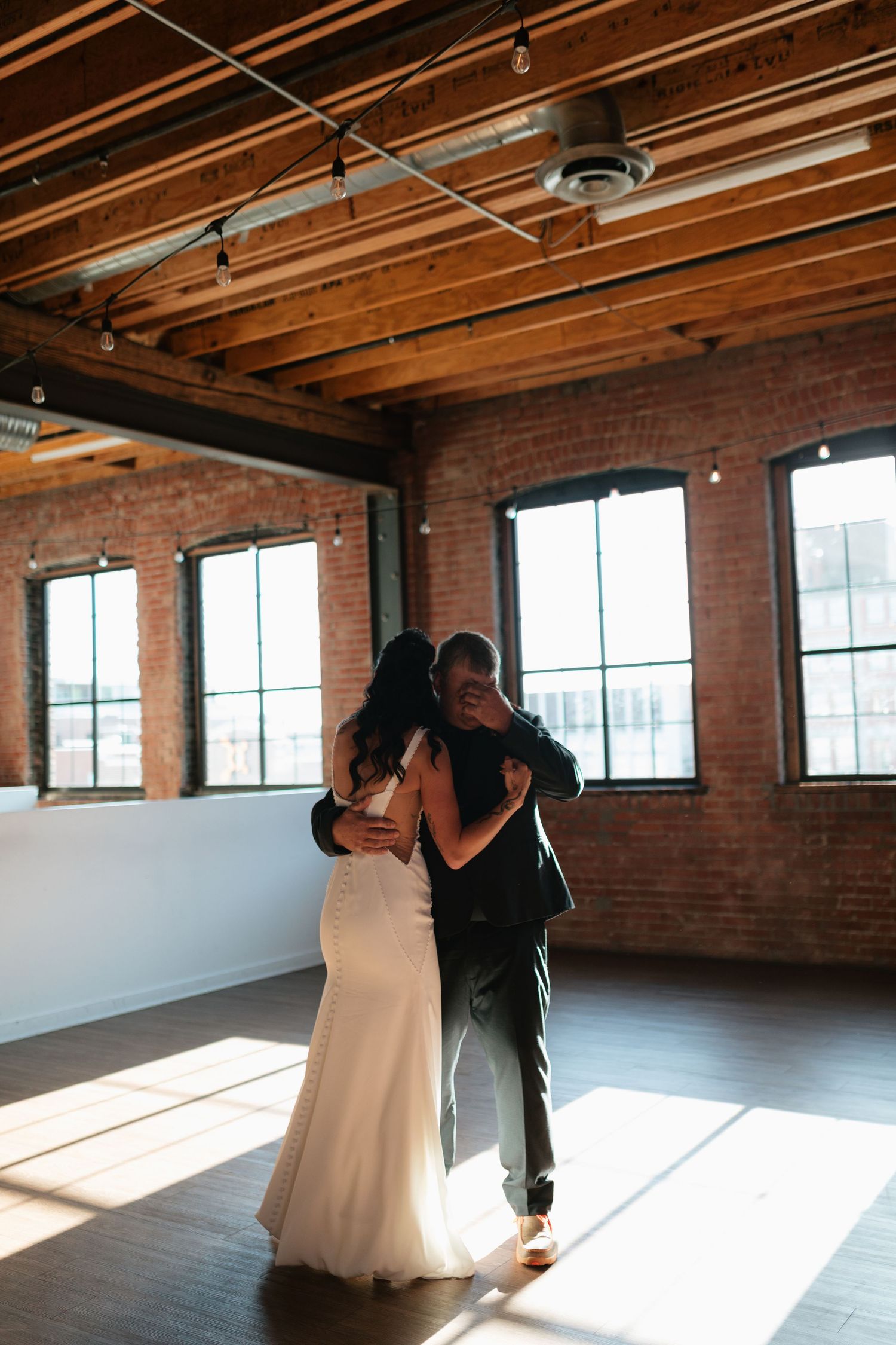A bride and groom share their first dance in a sunlit rustic venue with exposed brick walls and wooden beams.