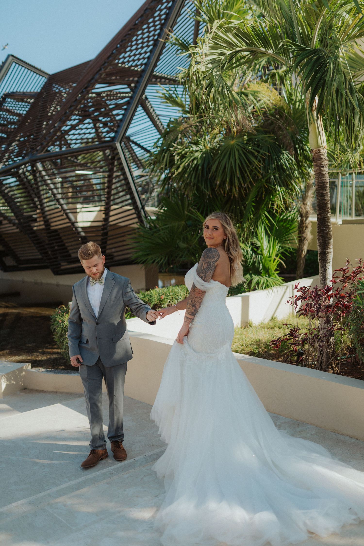 Bride in off-shoulder mermaid gown holding groom hand near tropical palm trees and resort structure.