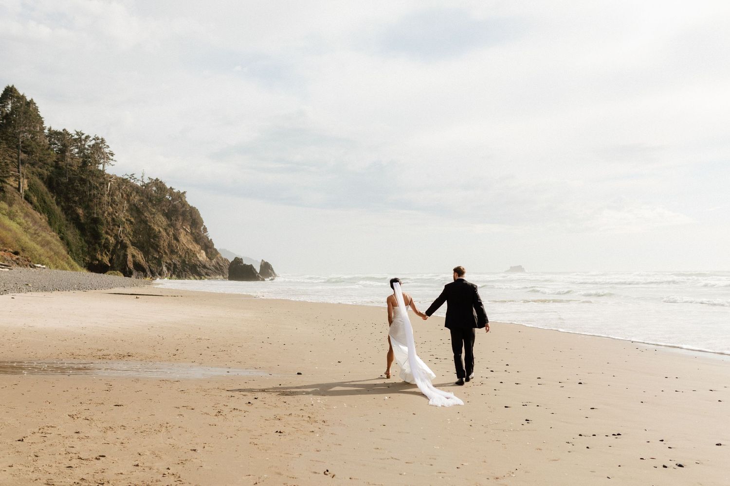 Bride and groom walk hand-in-hand along sandy beach shoreline with lush green cliffs in the background.