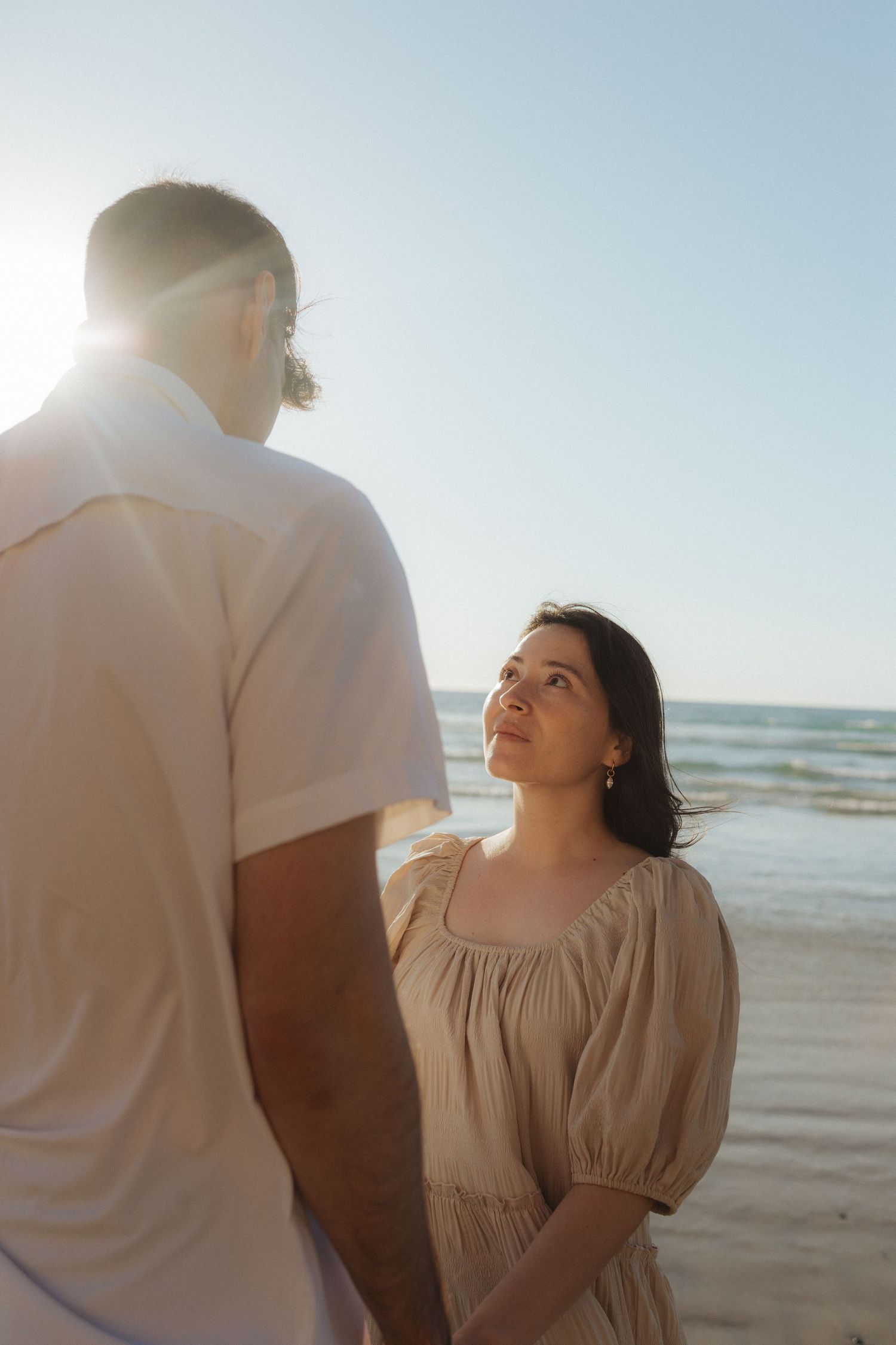Woman in beige dress gazes up at man in white shirt while holding hands on a sunlit ocean beach.