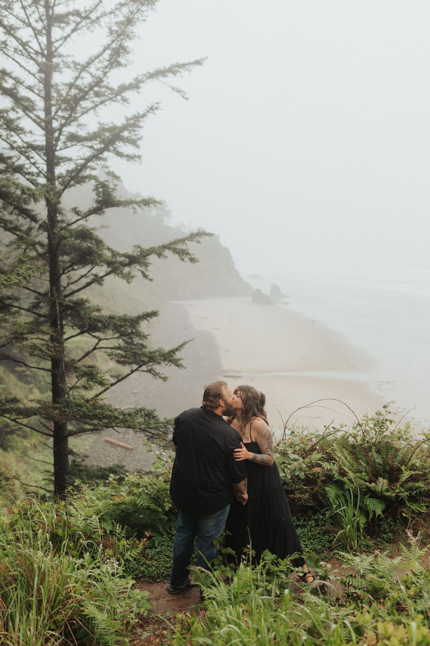 Couple sharing a kiss among lush green ferns on misty coastal cliffside with ocean in background.