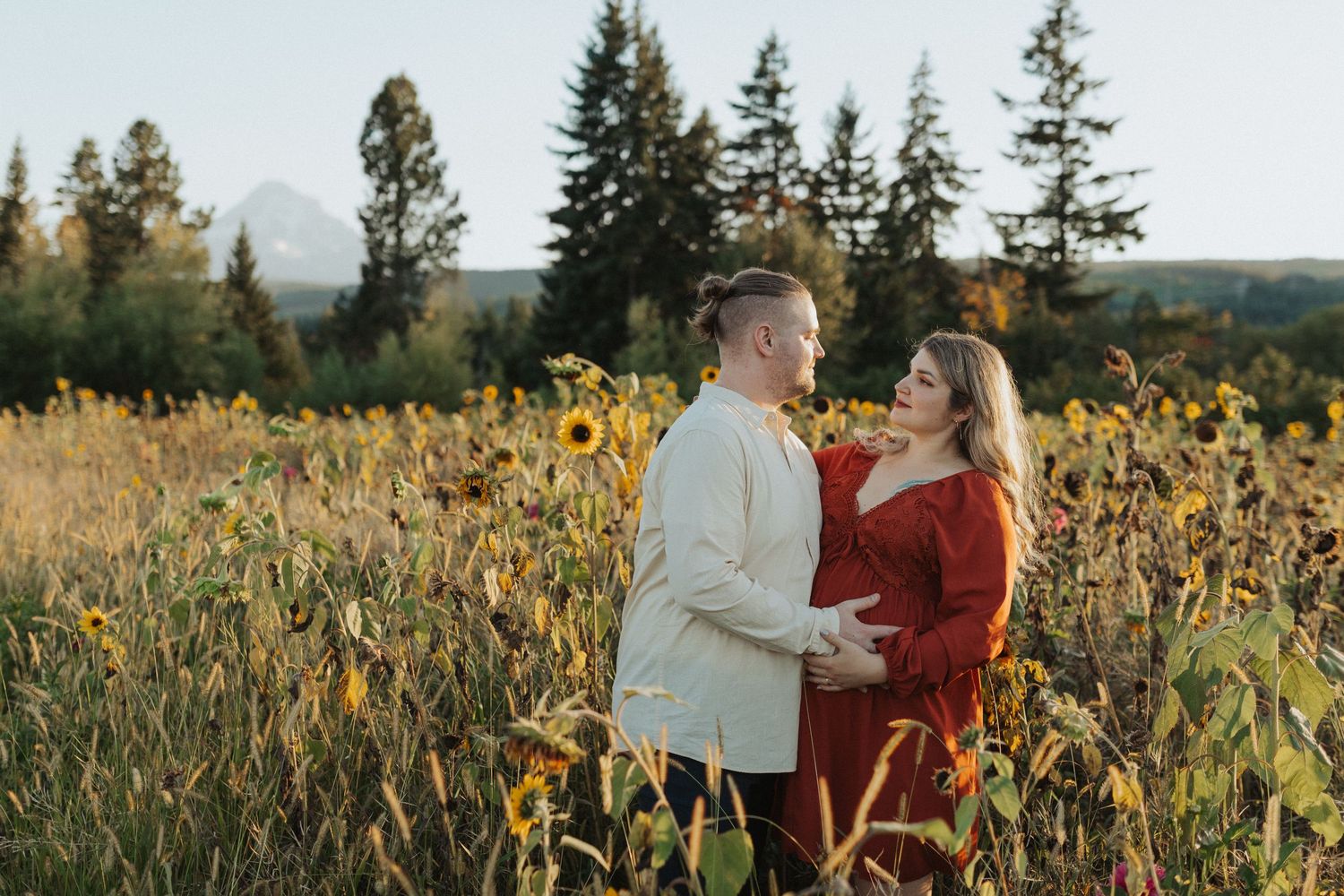 Expecting couple embracing in a sunflower field with a scenic mountain and evergreen tree backdrop.