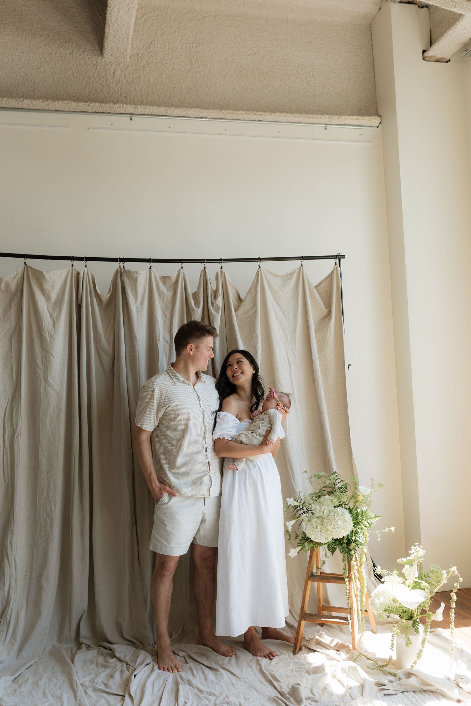 Young couple in neutral tones holding newborn baby in front of beige draped backdrop with white florals.