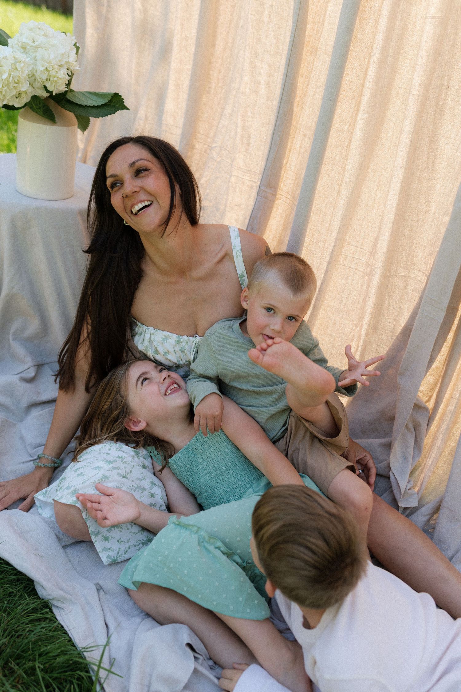 A mother in a floral dress holds her toddler while her son stands beside her in a sunny garden.