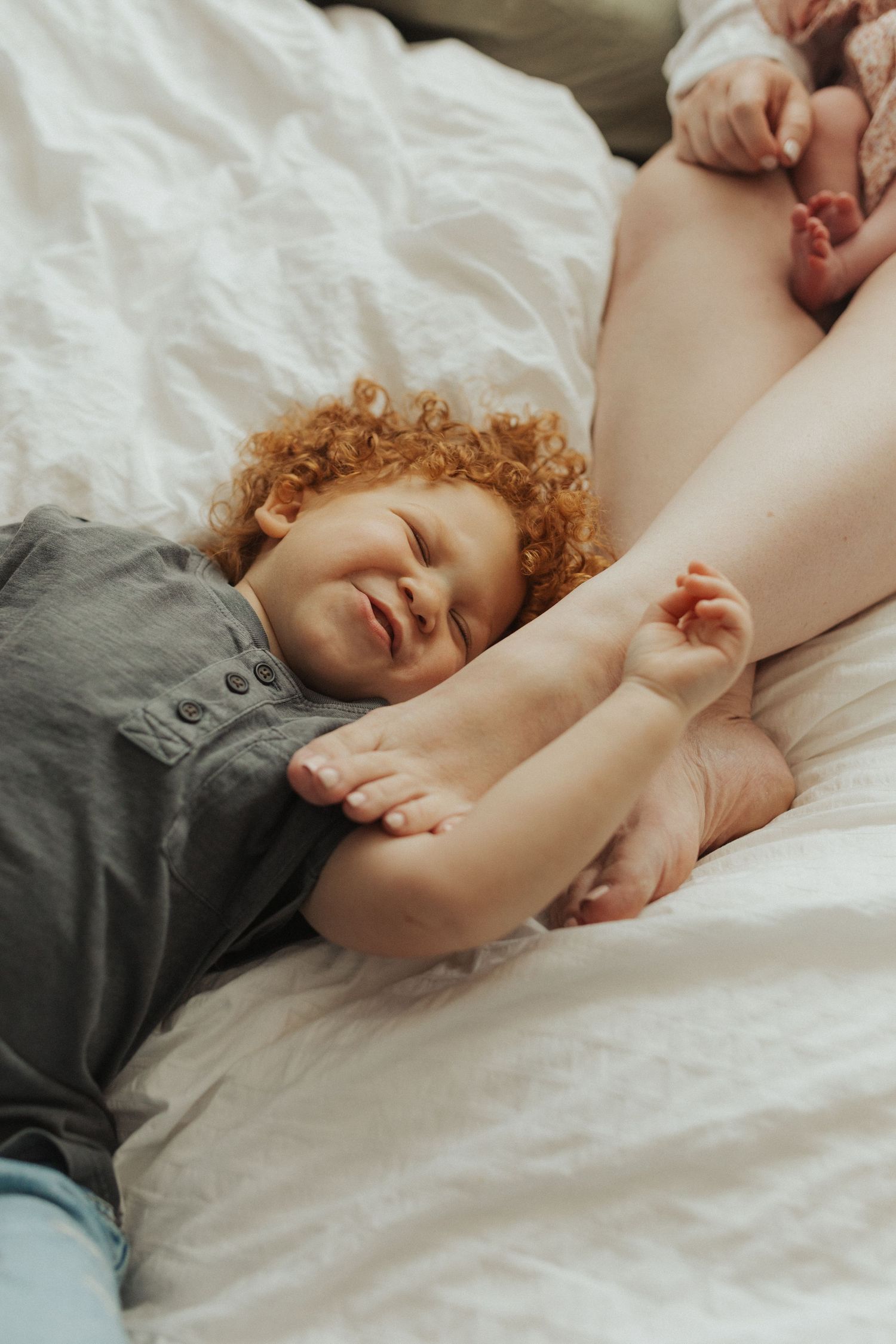 A curly red-haired toddler smiles while lying on a white bed next to a newborn baby.