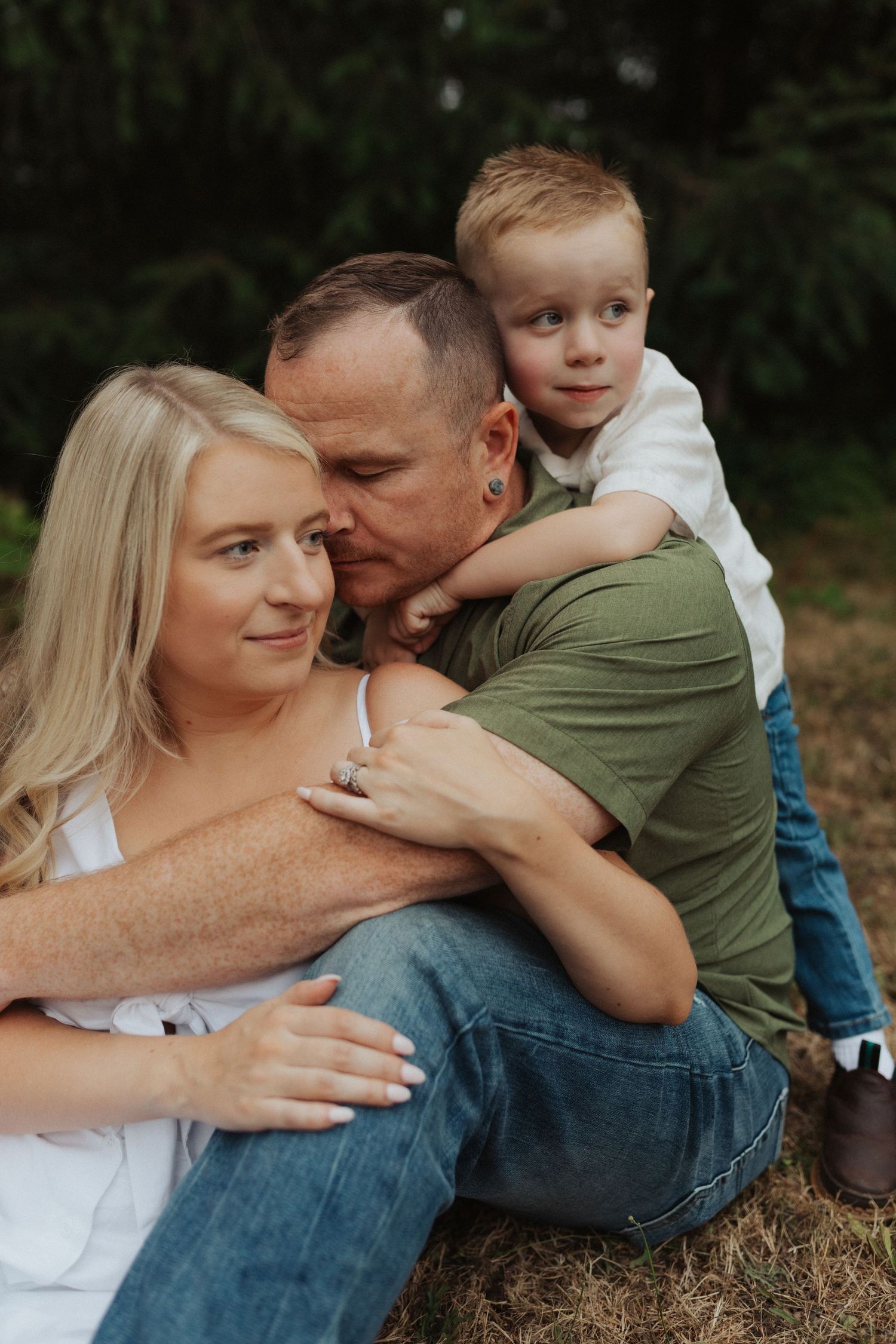 Family of three sitting together outdoors, with young boy hugging his dad from behind near trees.