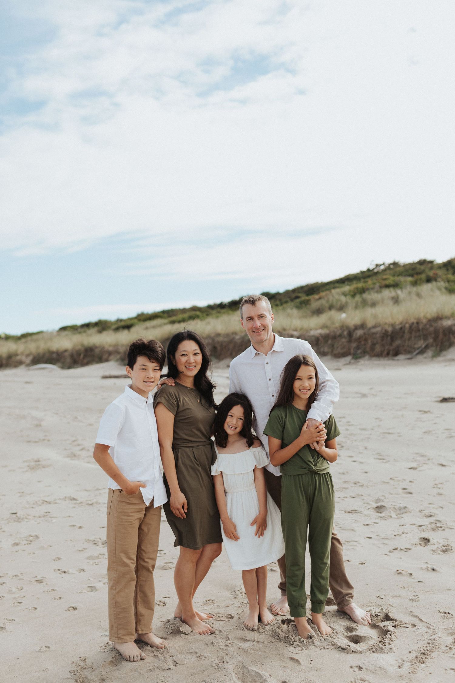 Family of five smiling together on a sandy beach, parents with three children in coordinated earth-tone outfits.