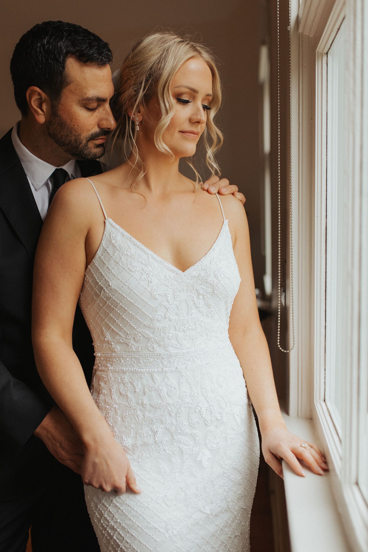 Blonde bride in beaded spaghetti-strap gown stands by window as groom in black suit embraces her from behind.