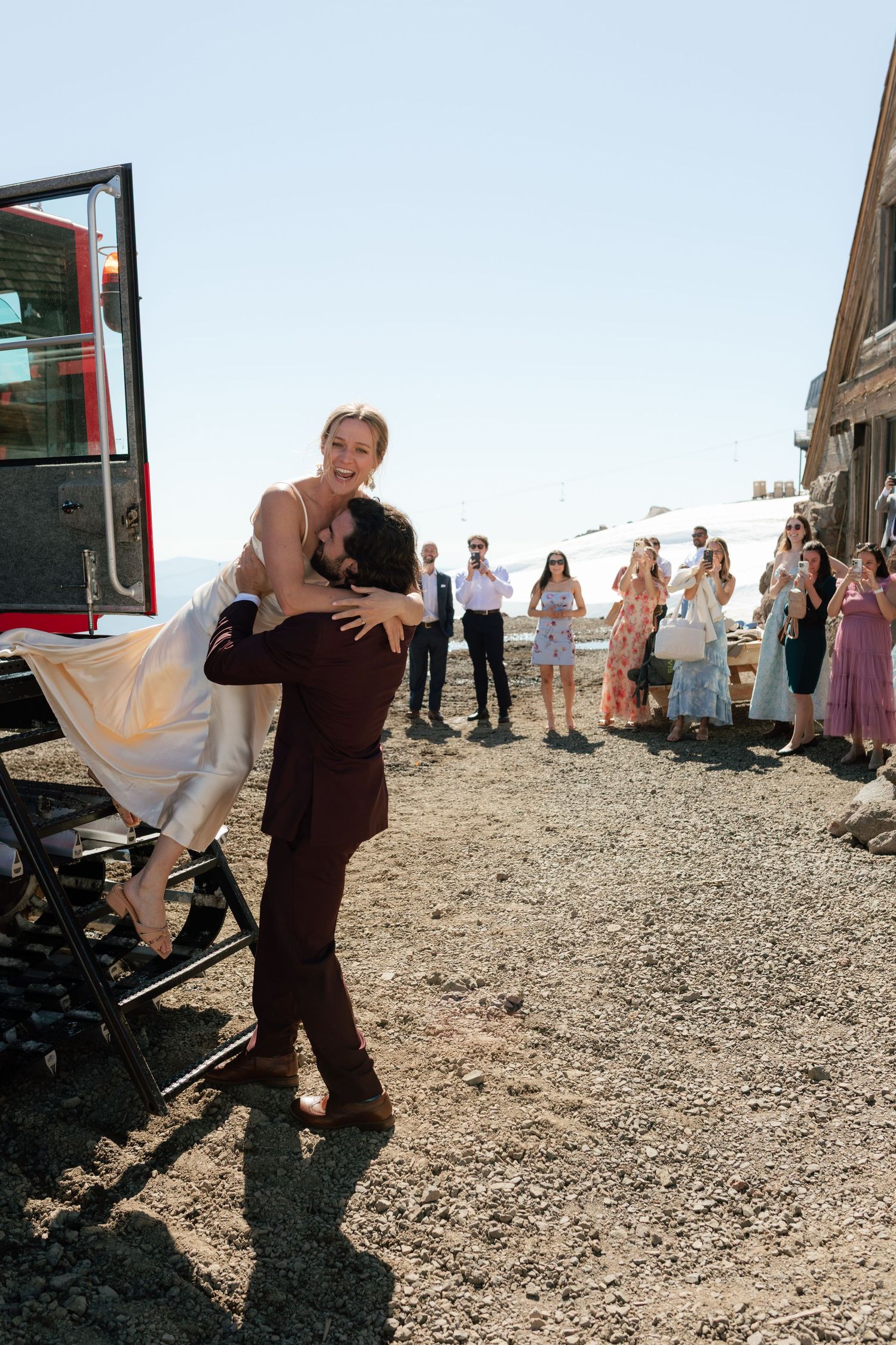 Groom lifts bride outside mountain wedding venue as guests celebrate nearby.