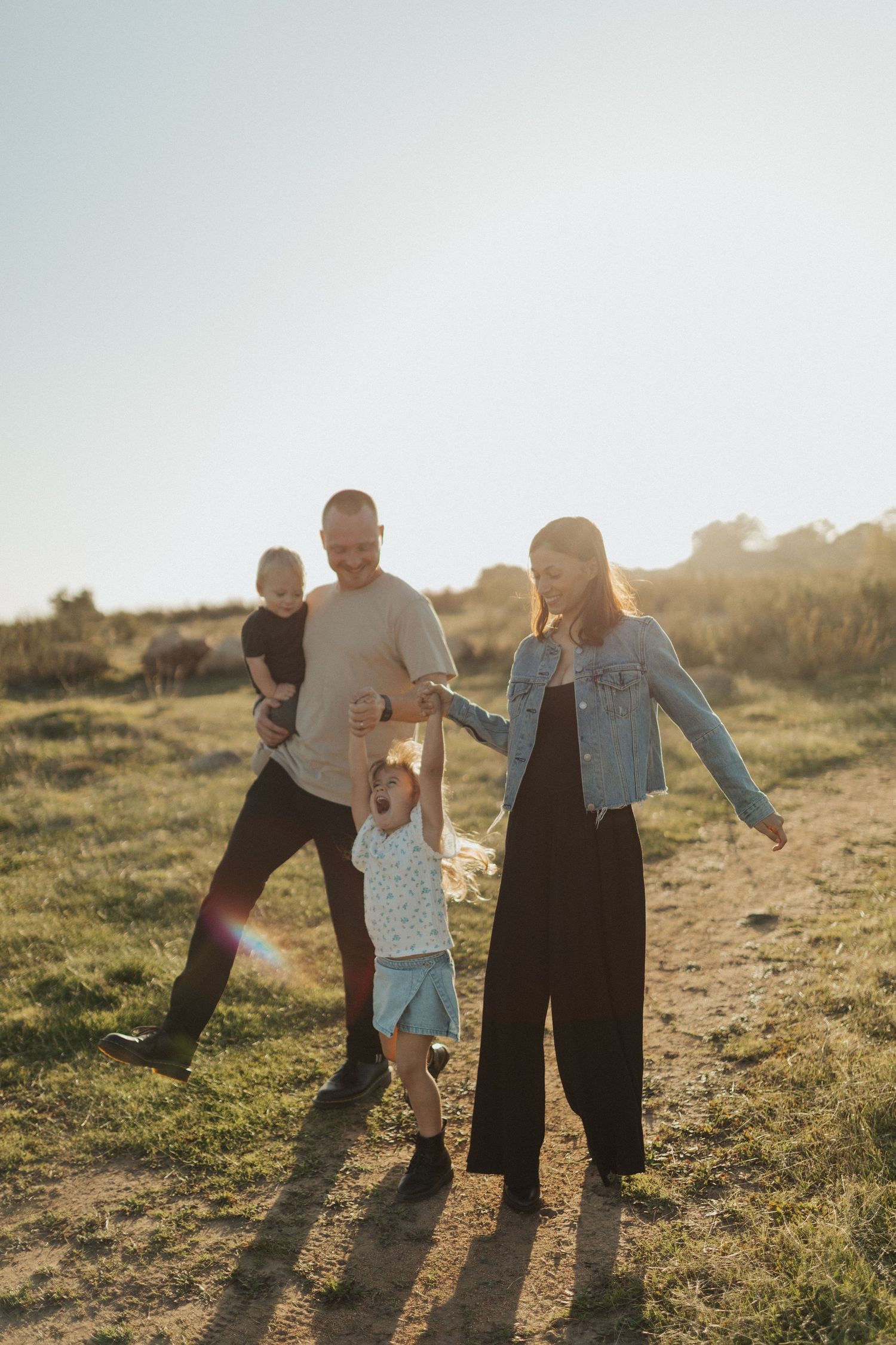 Joyful family of four playing outdoors in sunlit green field, parents swinging young daughter.