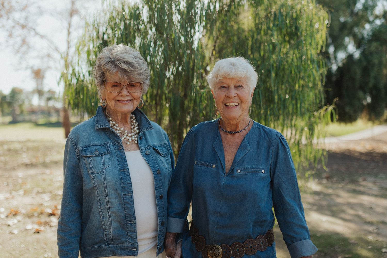 Two senior women with short white hair smile together outdoors near a weeping willow tree.