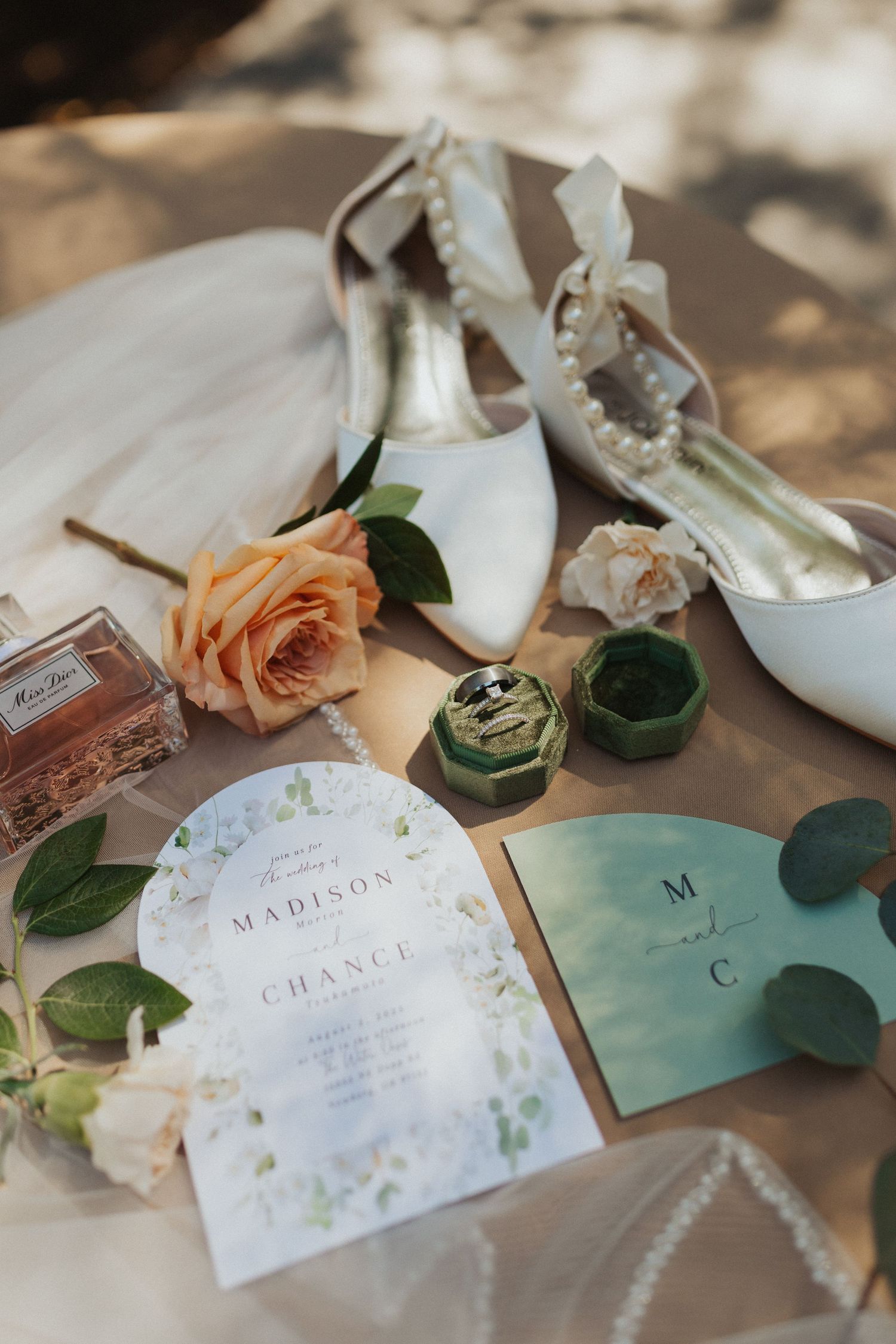 Elegant wedding flat lay featuring white heels, rings, floral invitations, and Miss Dior perfume on neutral fabric.