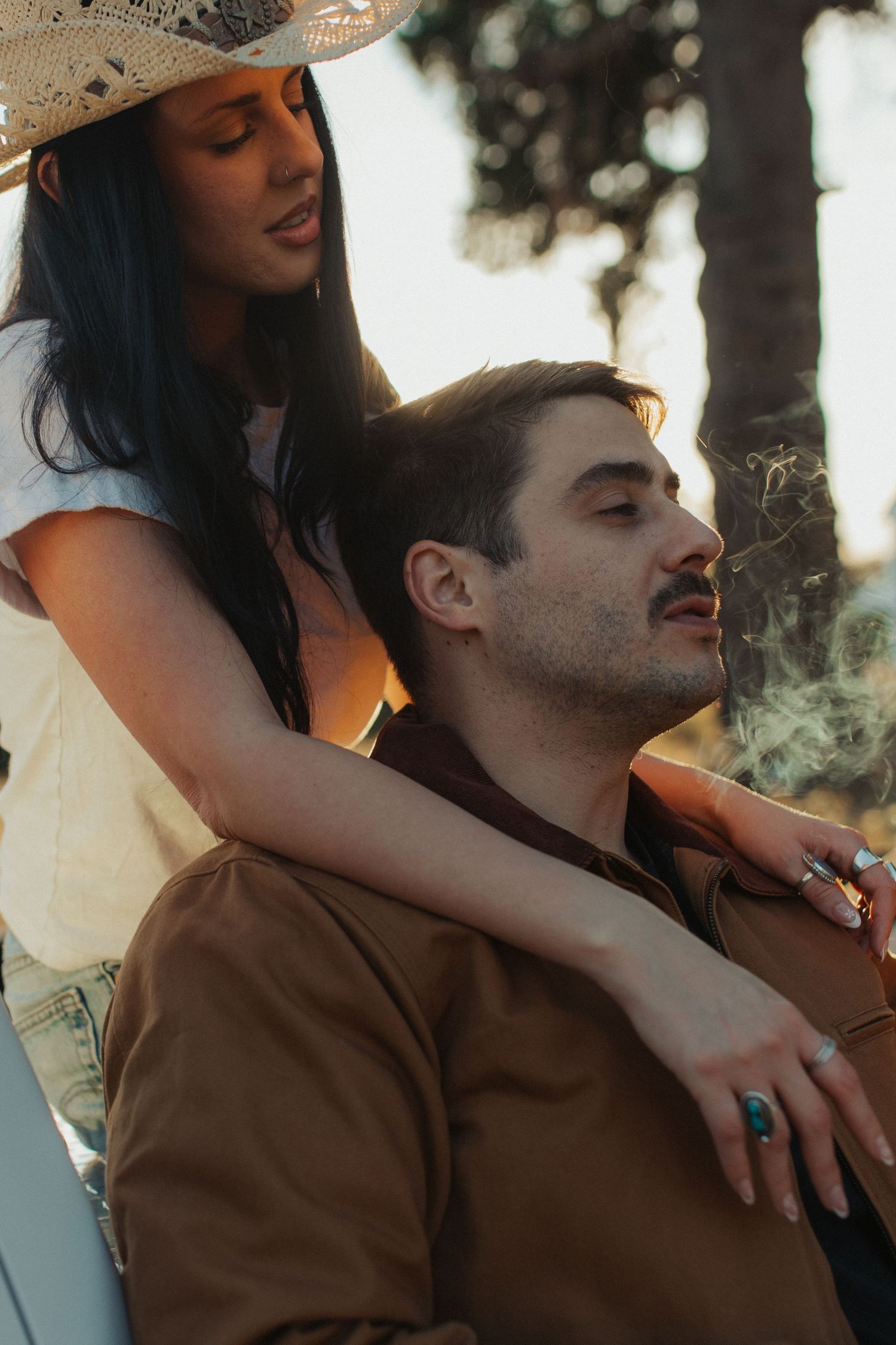 A woman in a cowboy hat embraces a man from behind as he smokes in warm golden light outdoors.