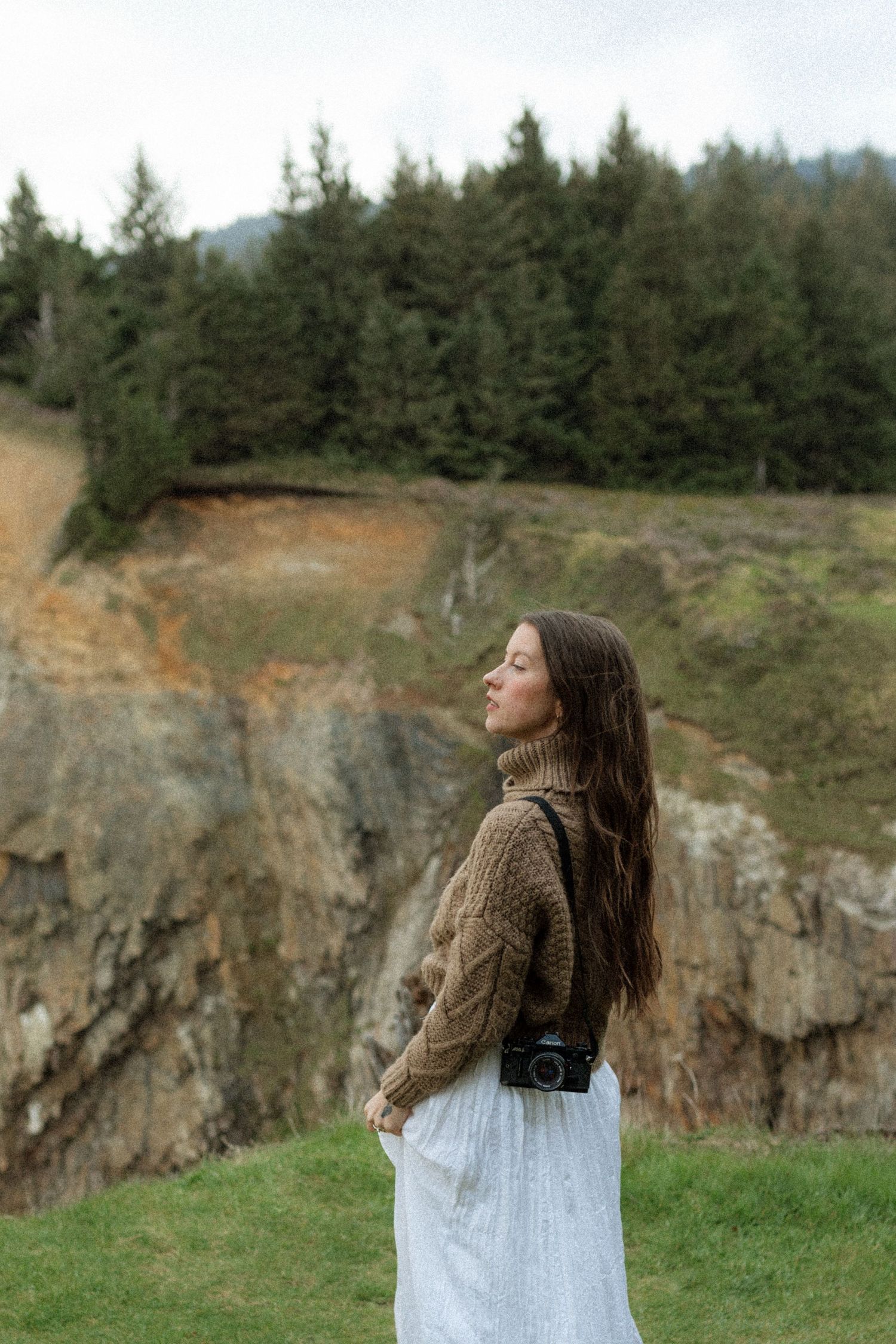 Woman in cable-knit sweater and white skirt standing before rocky cliffs and evergreen forest. Oregon wedding photographer.
