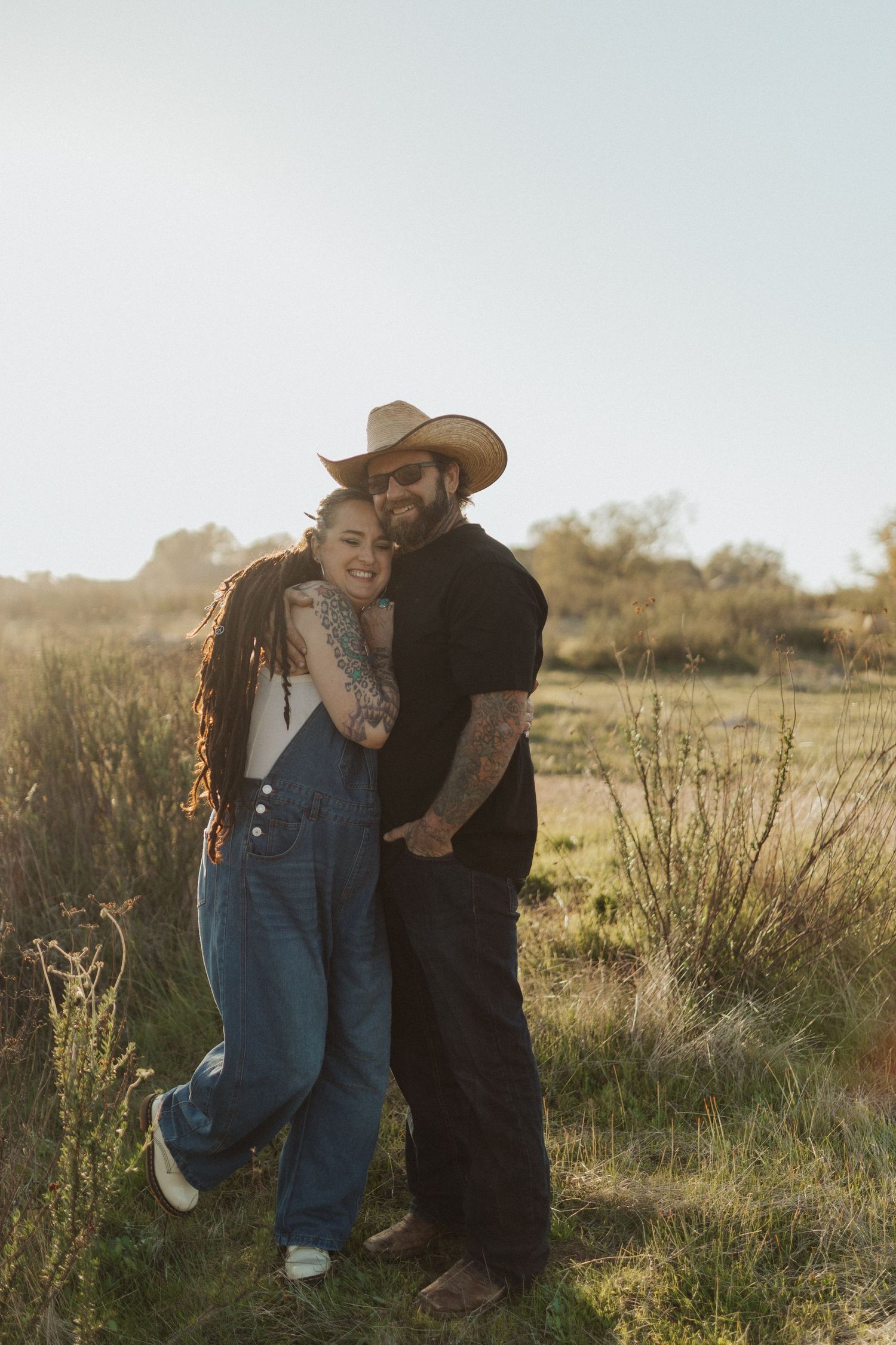 Tattooed couple embracing in a sunny field, man wearing cowboy hat, both smiling at camera.