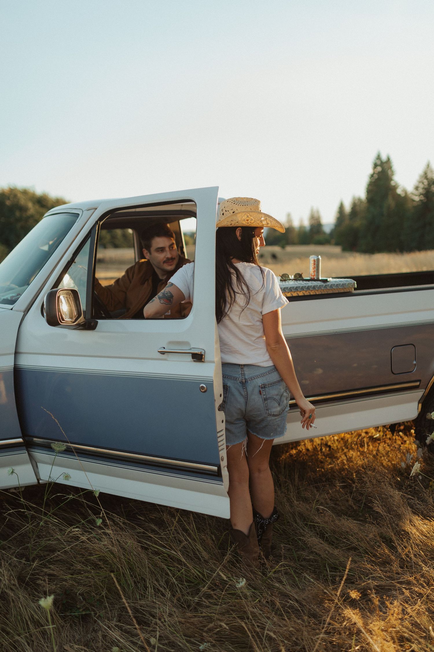 Woman in cowboy hat and denim shorts standing beside vintage pickup truck as man smiles from drivers seat.
