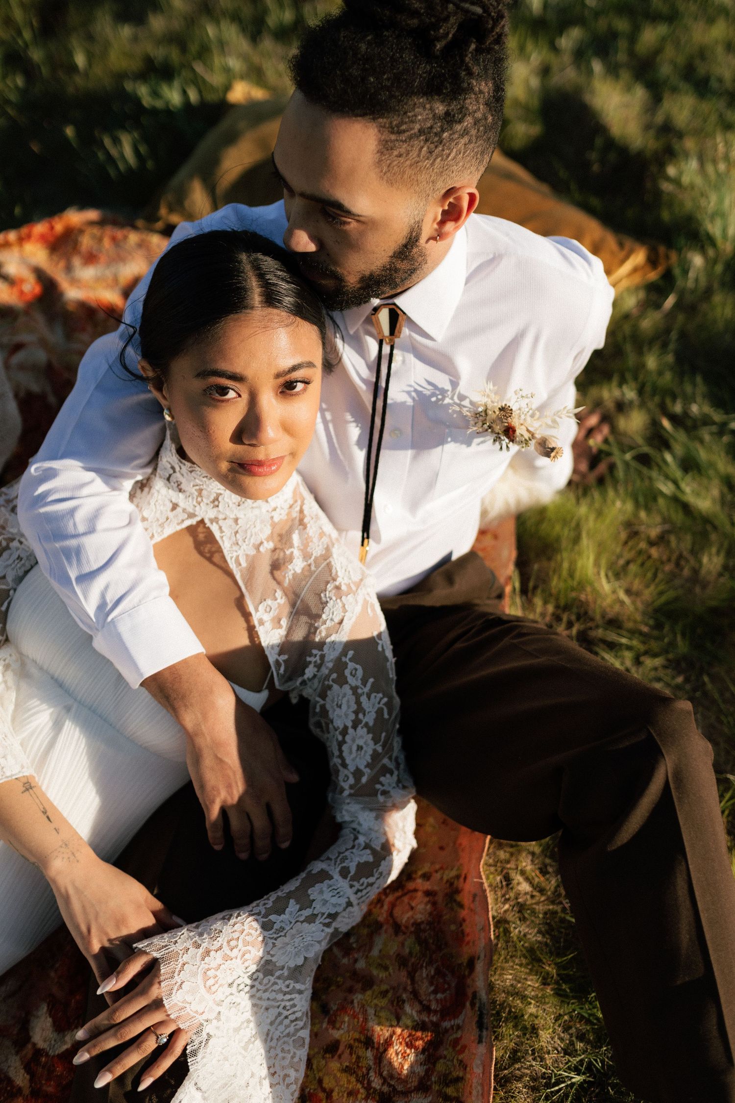 Groom tenderly embraces bride from behind during golden hour outdoor wedding shoot on a grassy hillside.