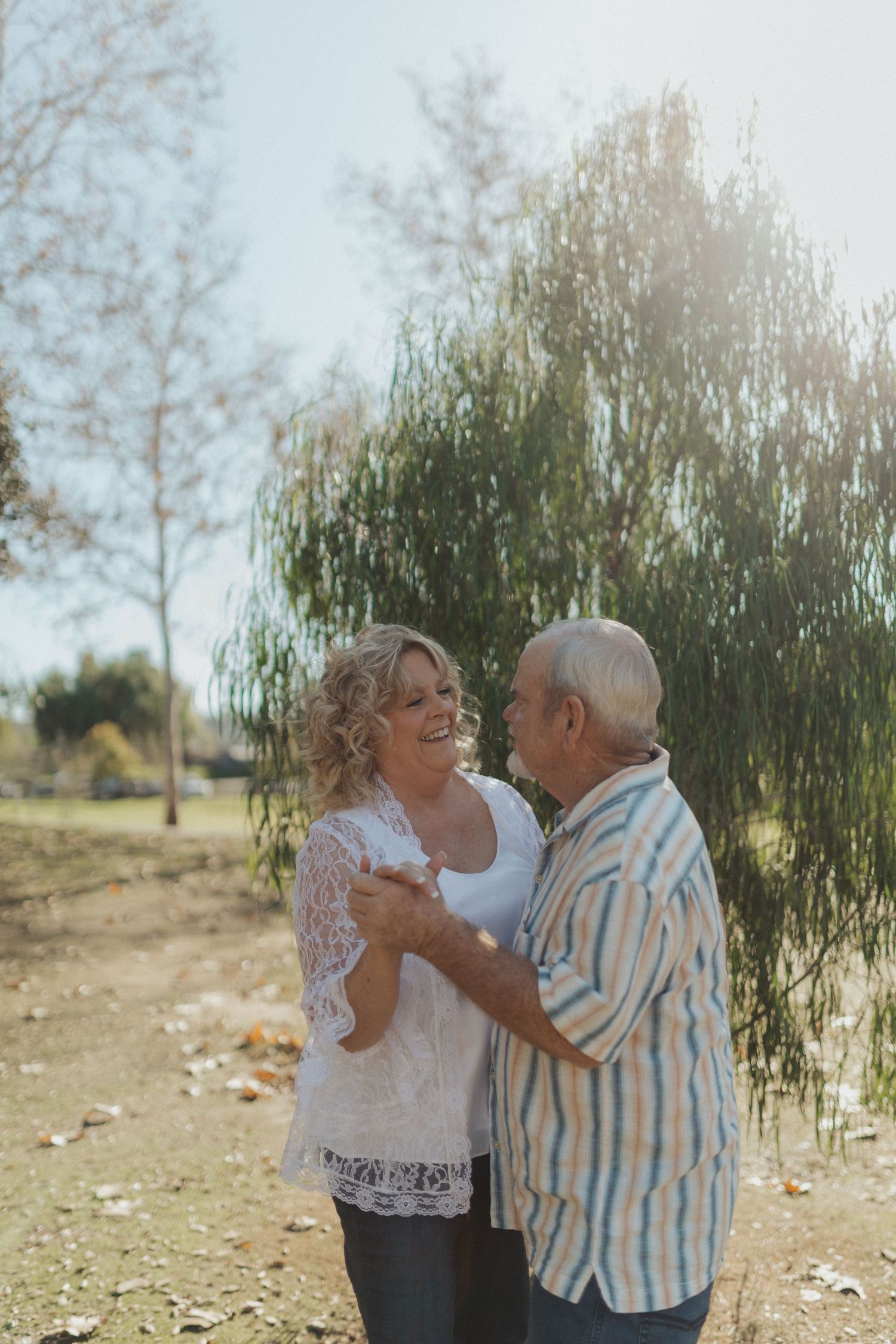 A joyful senior couple dances together in a sunlit park near a weeping willow tree.
