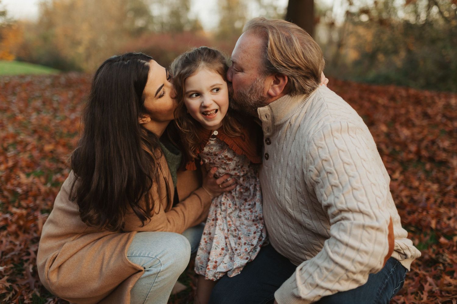 Two parents crouch down to kiss their smiling young daughter on the cheeks in an autumn setting.