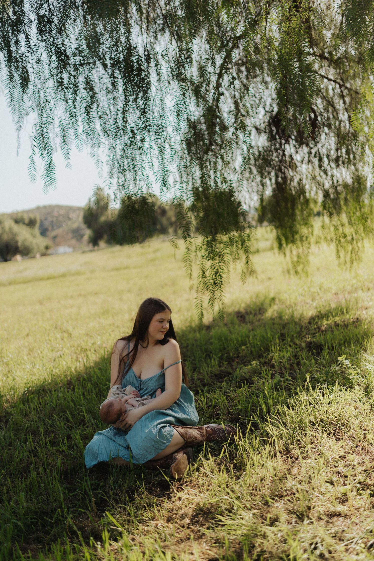 Mother in teal dress sitting in green grass nursing newborn under weeping willow tree outdoors.